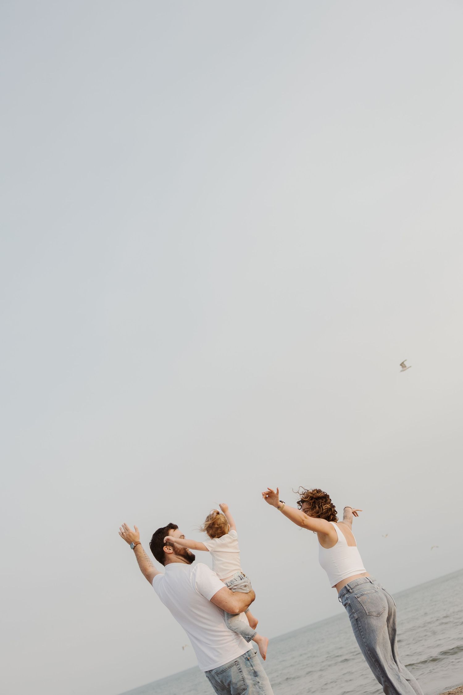 A family of three points at a bird in the sky while standing near the ocean.