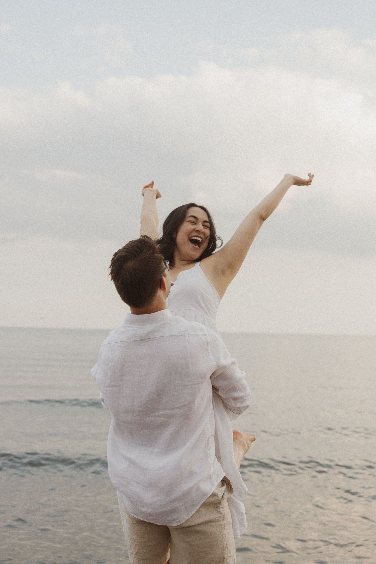 Man lifting joyful woman with arms raised near ocean at beach.