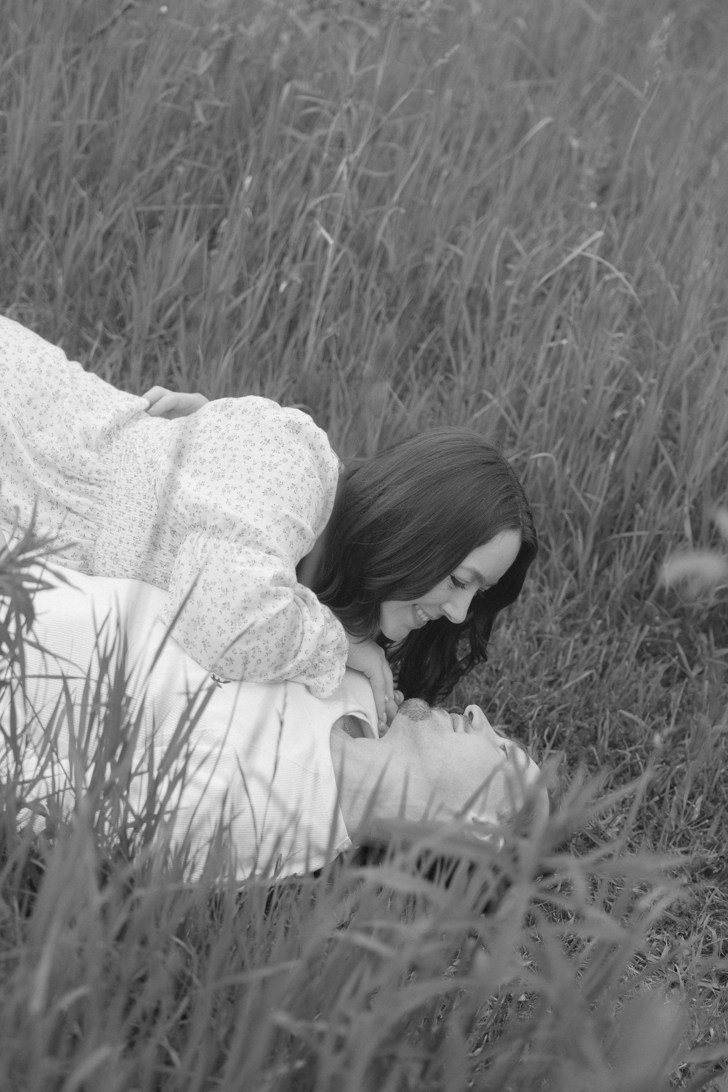 Black and white romantic photo of couple lying together in tall grass in an open field.