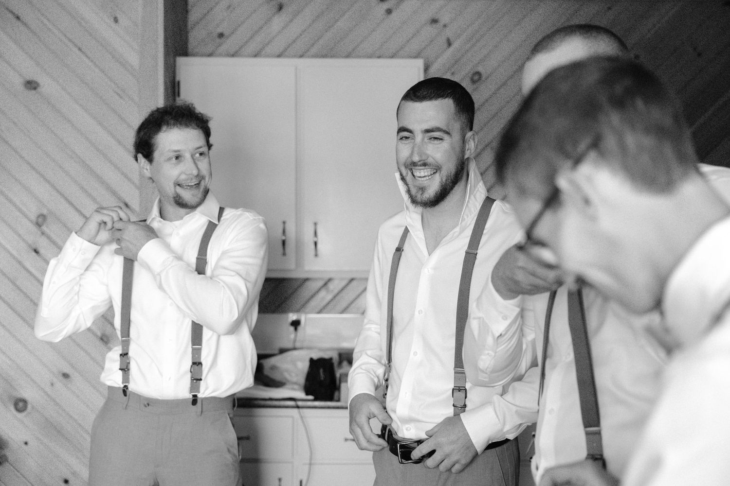 Black and white photo of groomsmen in suspenders laughing and getting ready together before the wedding ceremony.
