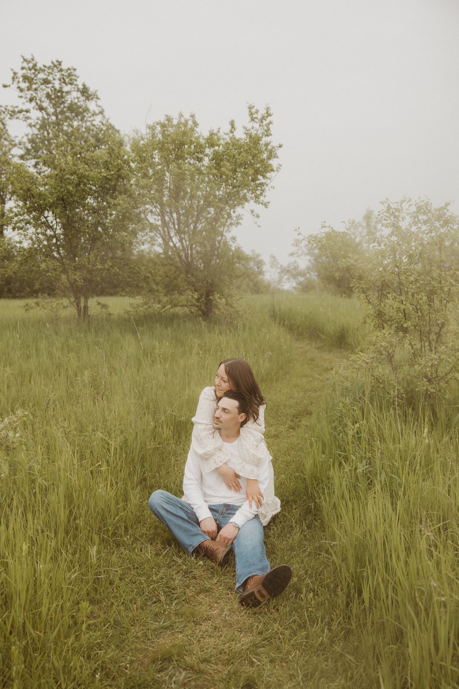 Couple sitting in tall green grass as woman embraces man from behind on a foggy day.