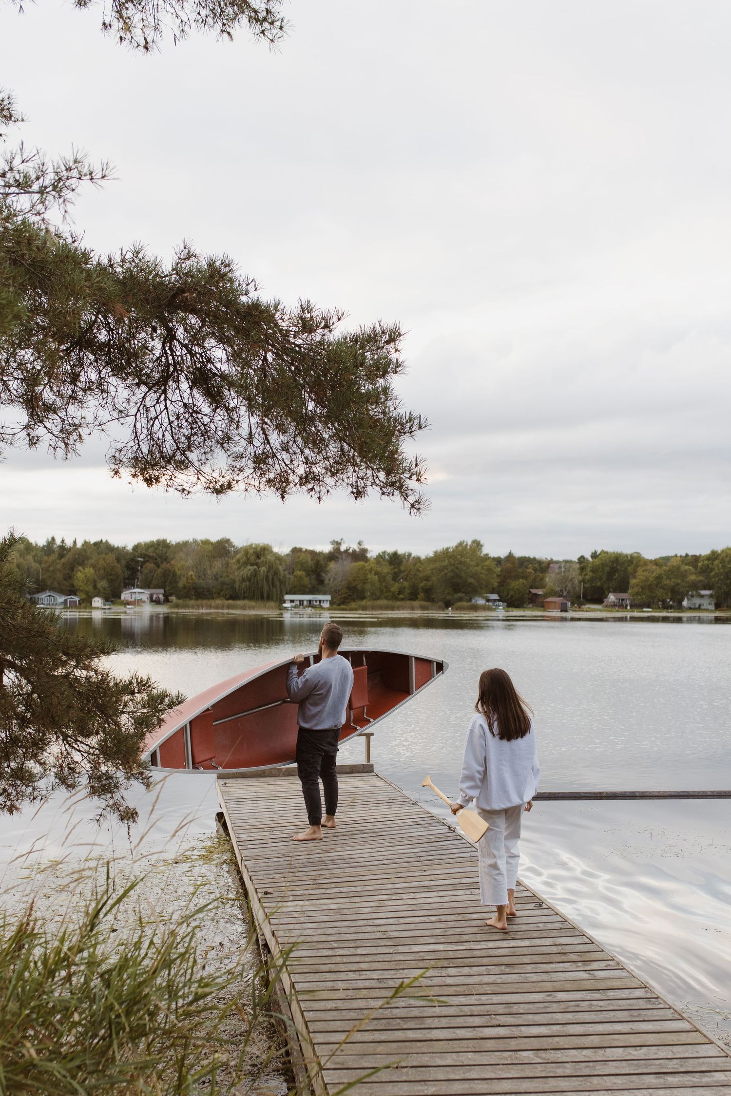 Two people walk barefoot on a wooden dock beside a red canoe on a calm lake surrounded by trees.