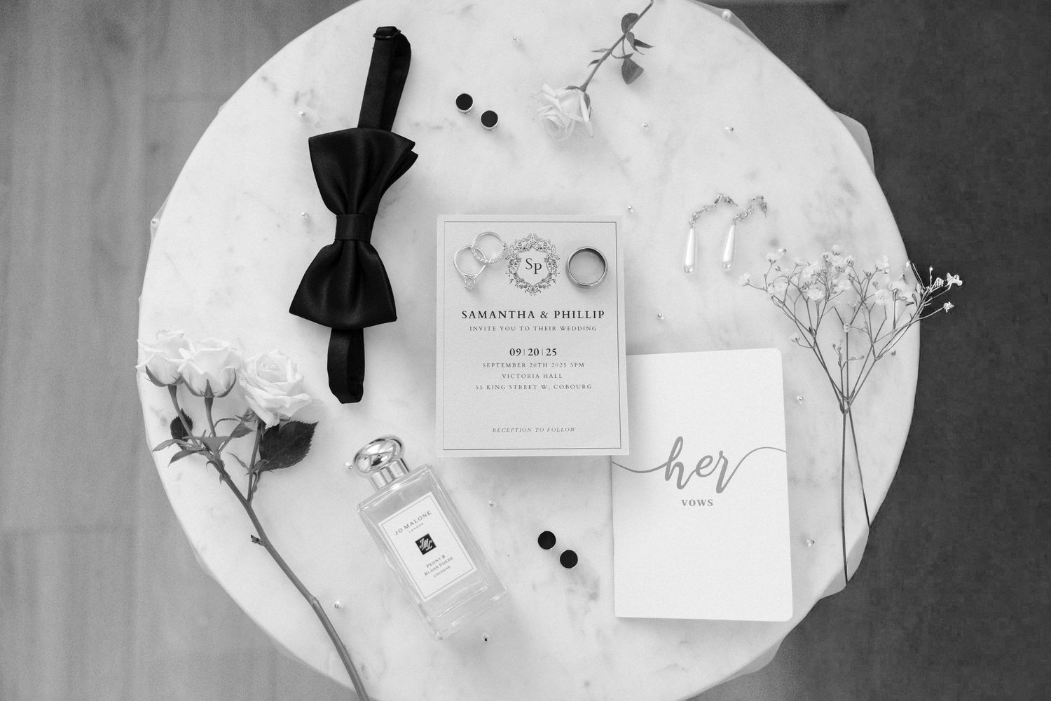 Black and white flat lay of wedding details including rings, bow tie, perfume, and invitation suite.
