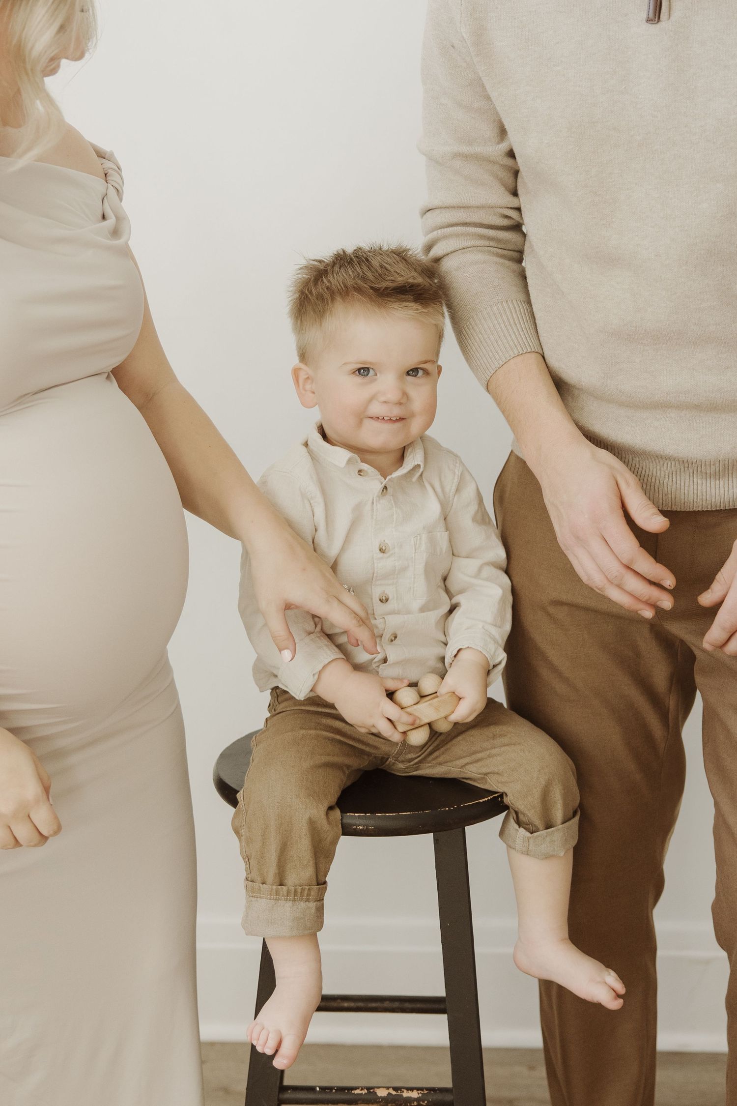Young boy sitting on a stool between his pregnant mother and father in a bright studio setting.