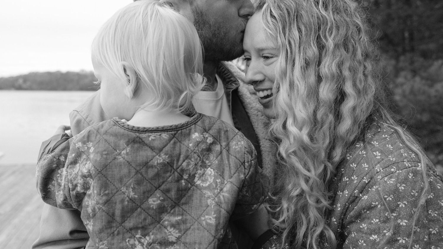 Black and white photo of parents sharing a tender moment with their young child by a lake.