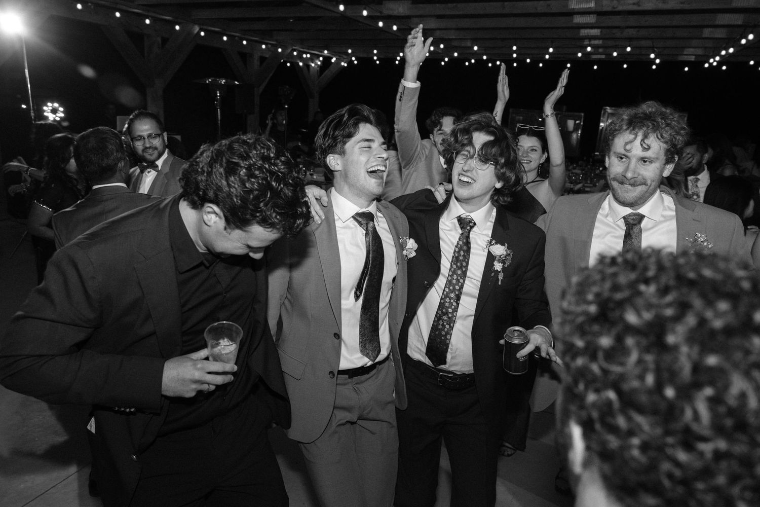 Black and white photo of groomsmen and guests laughing and dancing together at a lively wedding reception.