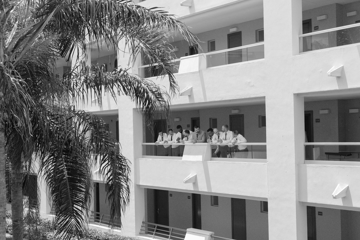 Black and white photo of people standing on balconies of a multi-story tropical hotel building with palm trees.