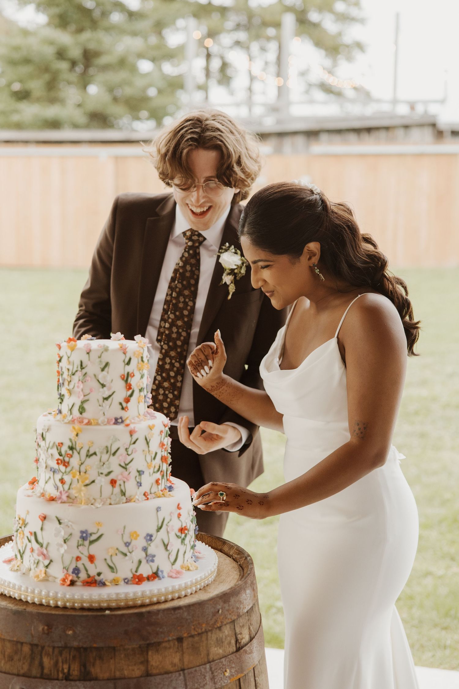 Bride and groom smiling while cutting a three-tier floral wedding cake placed on a rustic wooden barrel outdoors.
