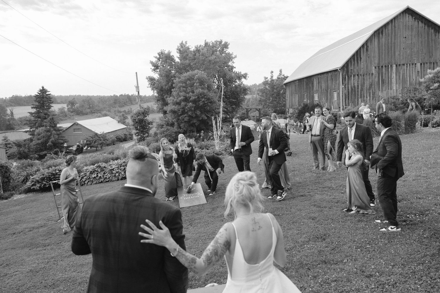 Black and white aerial view of bride and groom entering outdoor wedding reception near rustic barn.