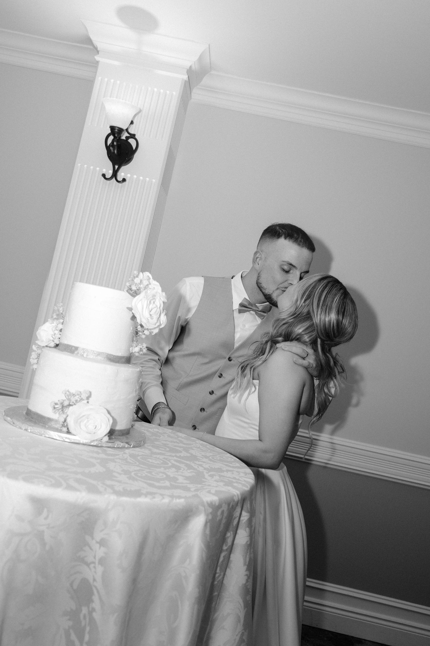 Black and white photo of bride and groom kissing while cutting their elegant three-tier wedding cake.