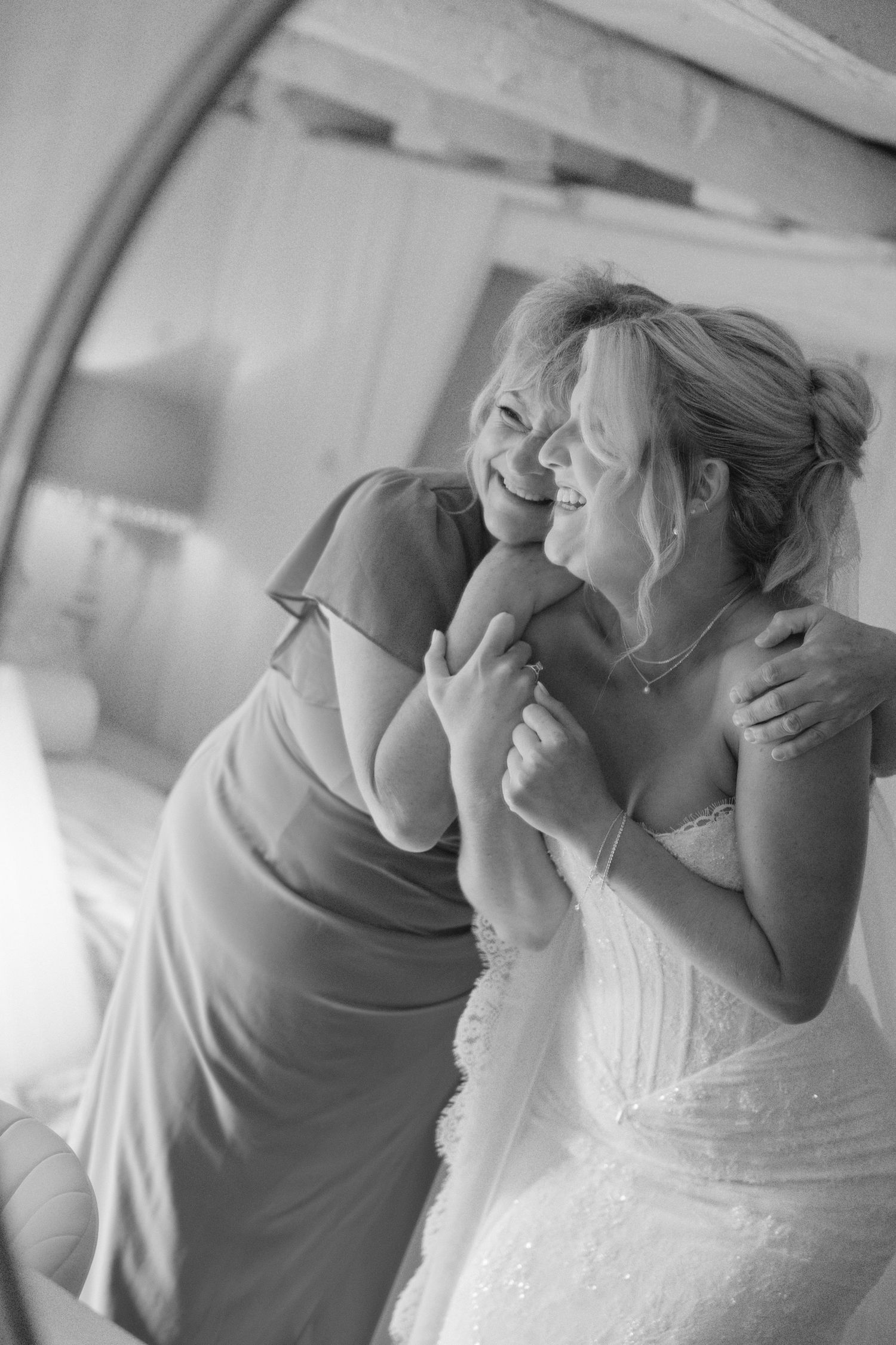 Black and white photo of bride in wedding dress laughing with woman in formal gown during bridal preparations.