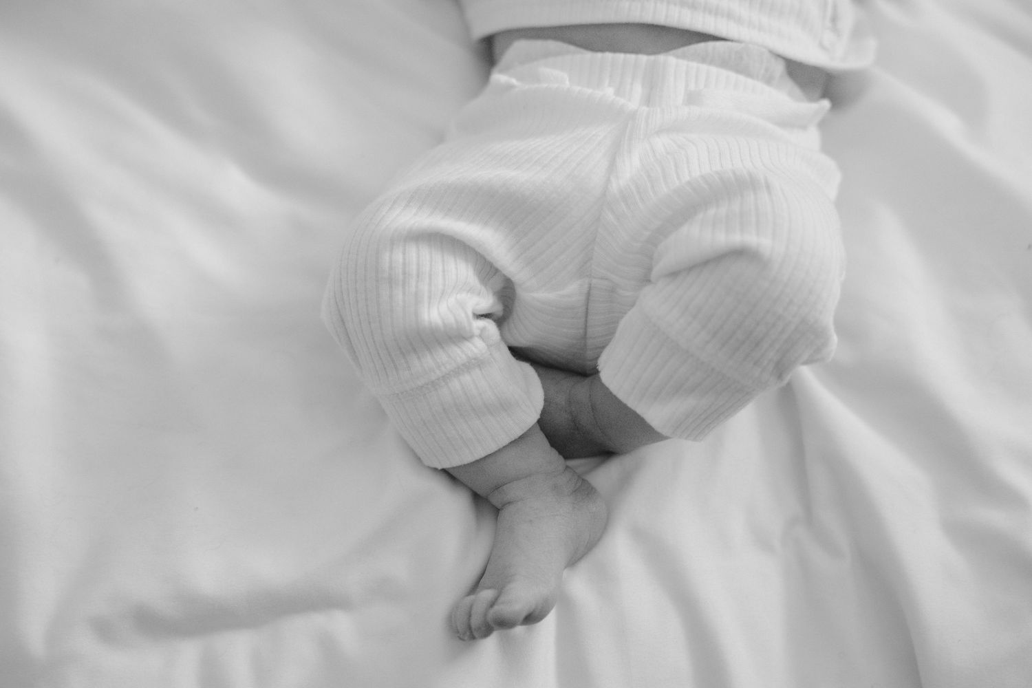 Black and white close-up of a sleeping newborn baby wearing white pants and socks on a soft white bed.