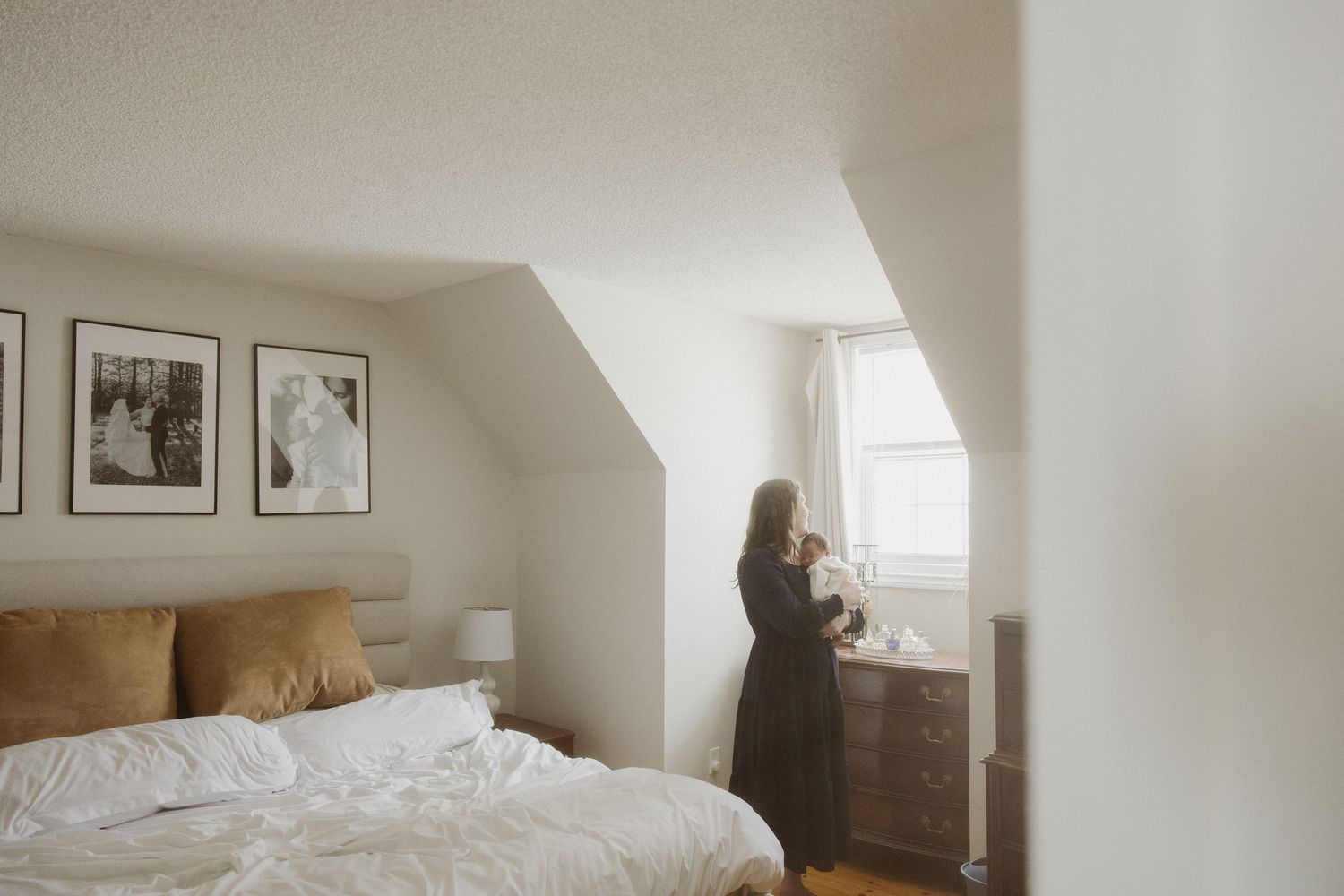 Woman standing by a sunlit window in a cozy bedroom with white bedding and framed wall art.