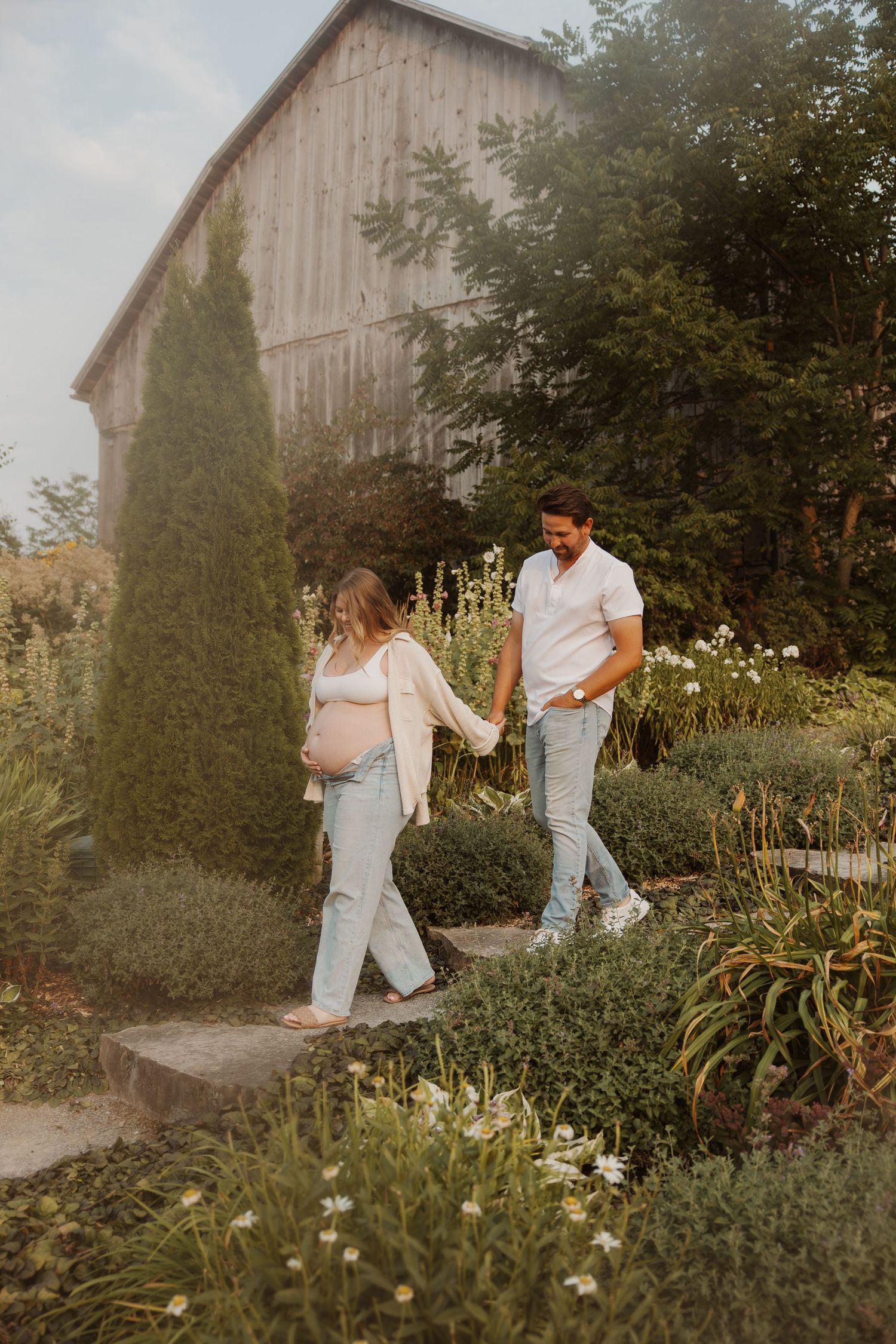 Pregnant woman and man walking hand in hand through a lush garden near a barn.