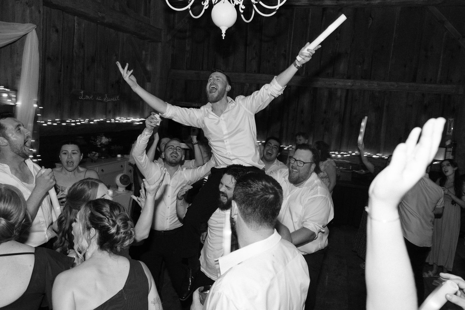 Excited wedding guest lifted on shoulders holding glow stick during lively barn reception dance party.