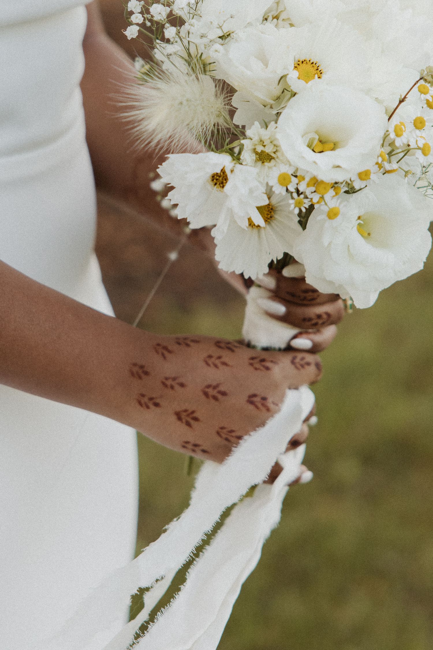 Close-up of bride holding a white floral bouquet with henna-adorned hands during an outdoor wedding.