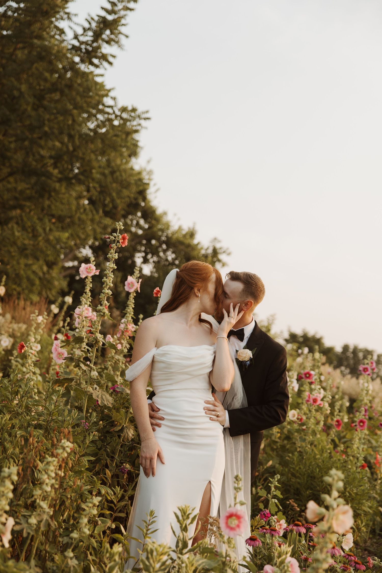 Bride and groom share a kiss surrounded by colorful wildflowers in a romantic garden setting.