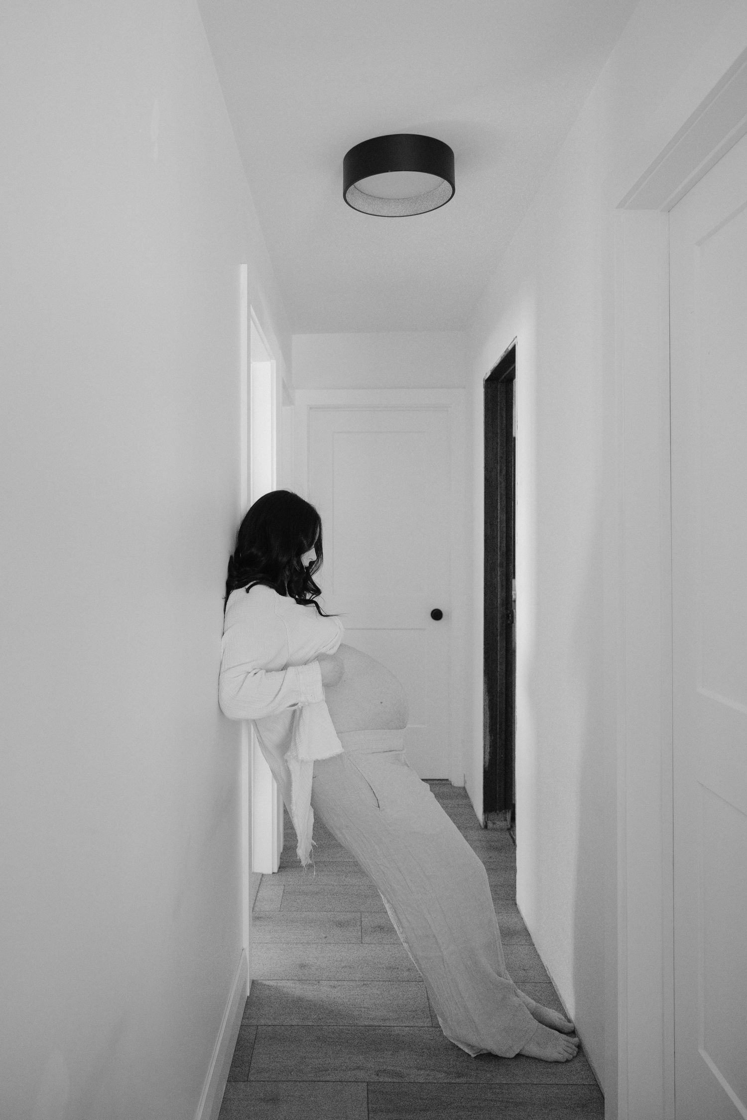 Black and white photo of pregnant woman cradling her baby bump in a bright white hallway.