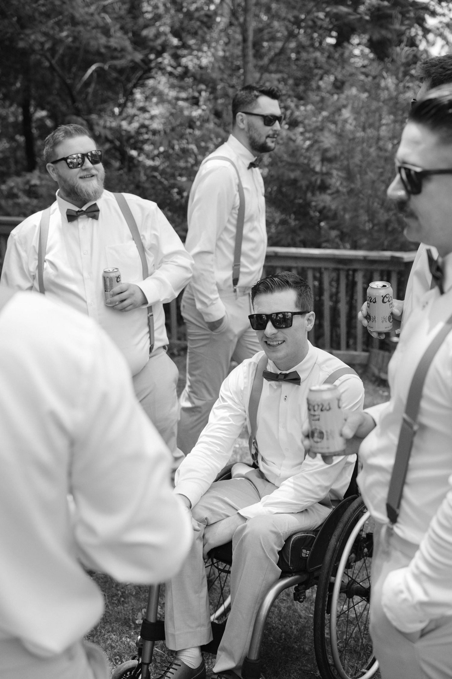 Black and white photo of groomsmen in suspenders and bow ties celebrating with drinks outdoors.