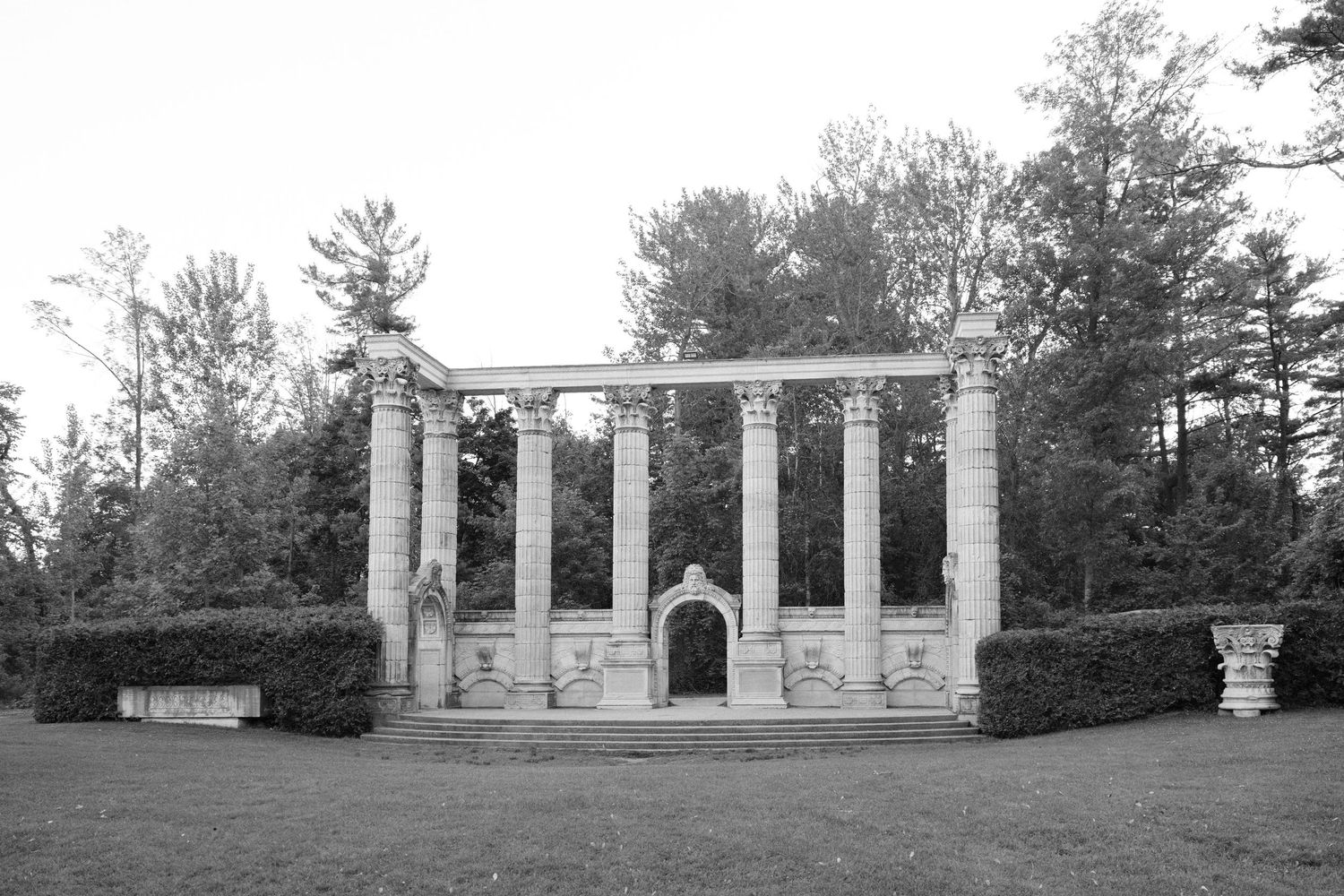 Black and white photo of classical stone columns and archway garden structure surrounded by trees.