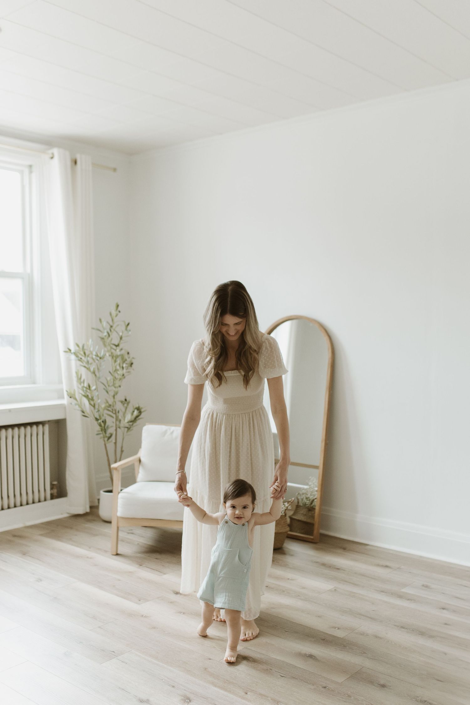 Young mother in cream dress holding hands with her toddler baby in a bright minimalist white room.