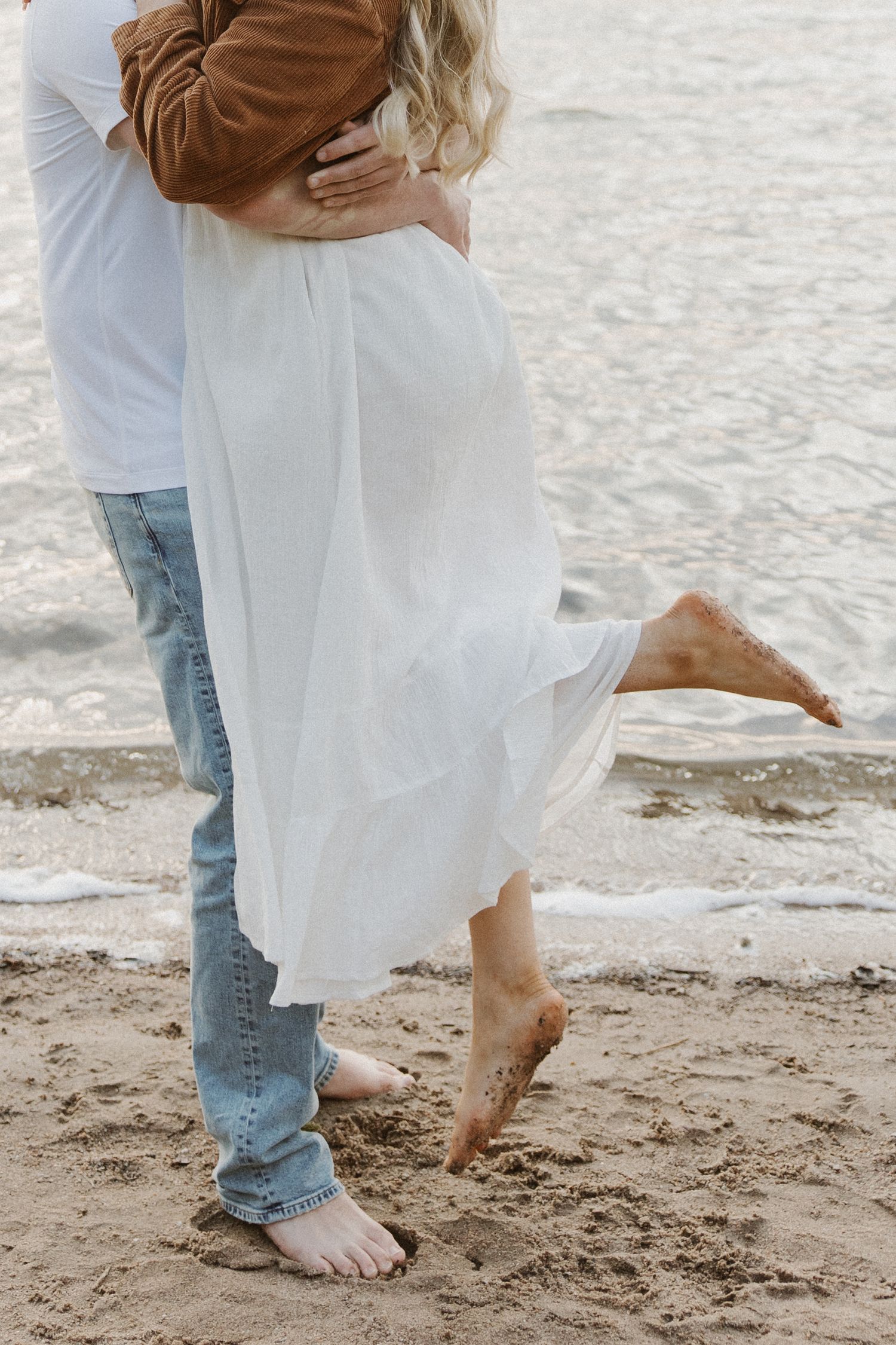 Barefoot couple embracing on sandy beach with ocean waves behind them.