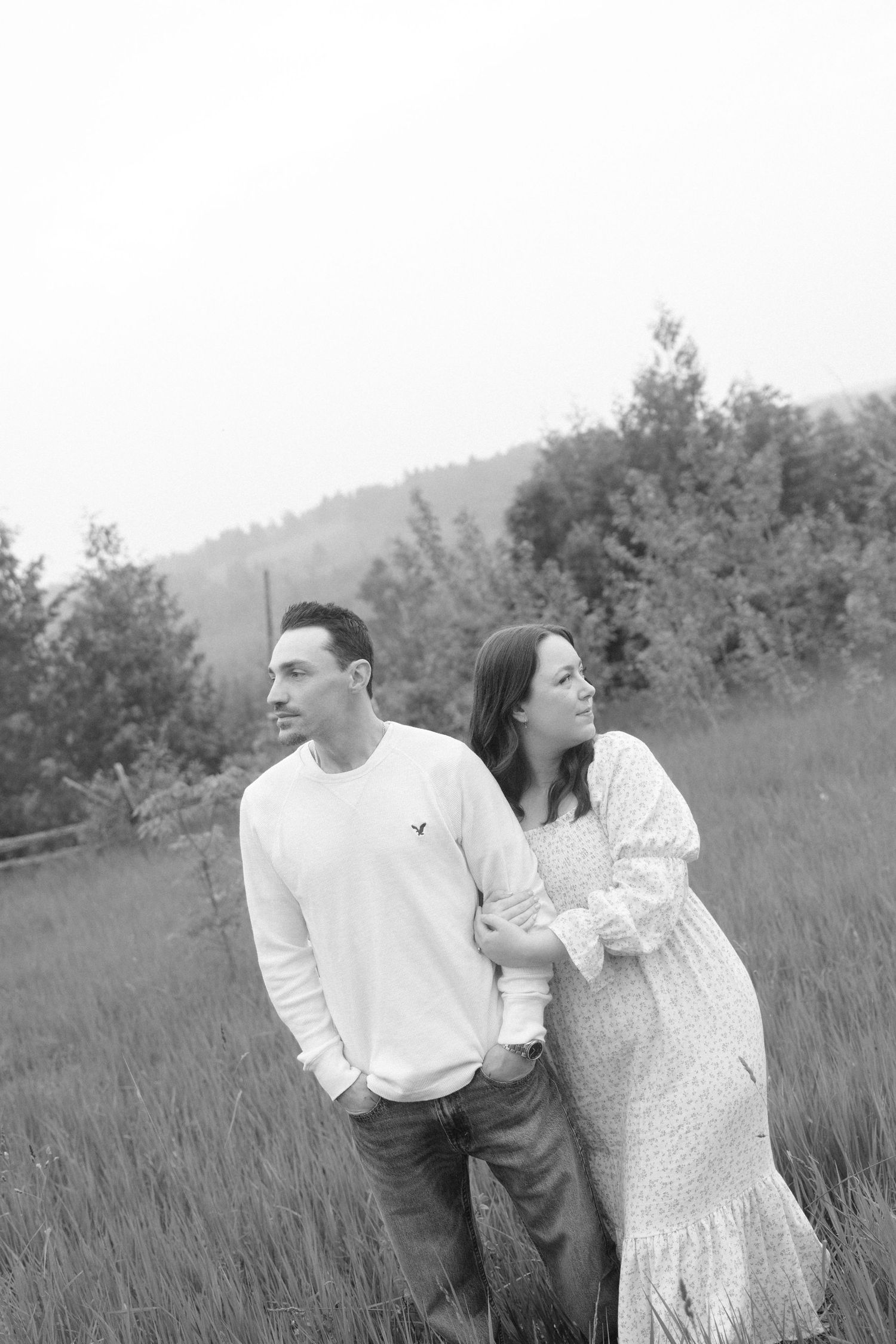 Black and white photo of couple standing together in a misty meadow with trees and hills behind them.