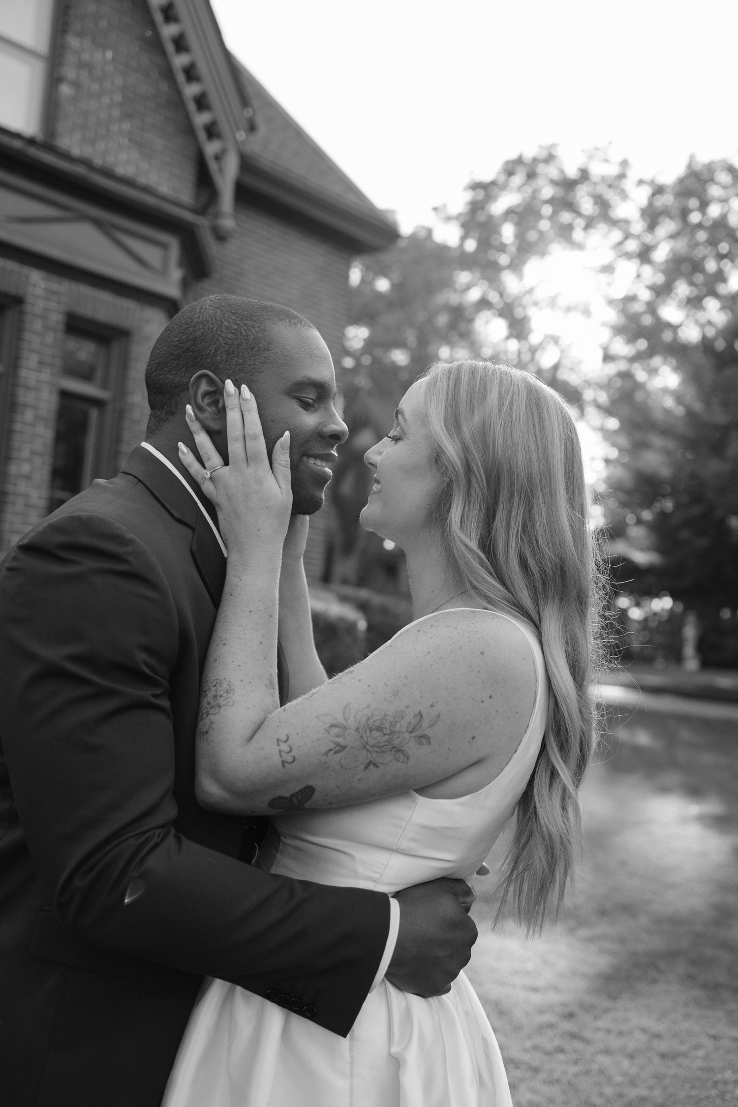 Black and white romantic portrait of interracial couple nearly kissing outside a historic brick building.
