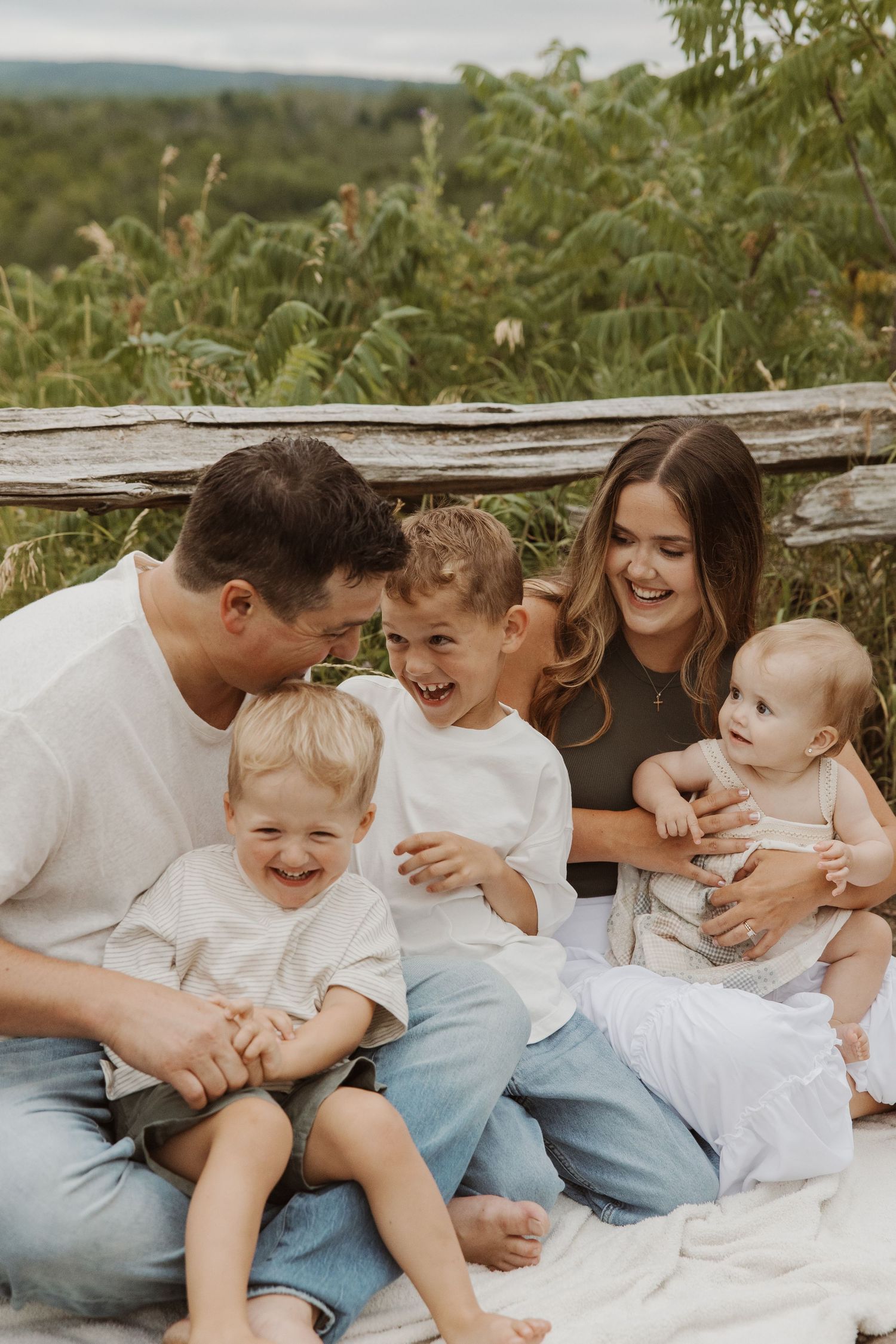 Happy family of five sitting together laughing on a blanket in a natural setting.