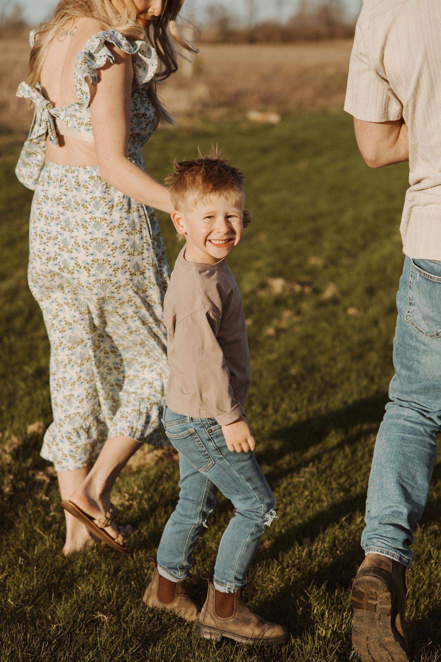 Happy red-haired toddler boy smiling while walking outdoors with his parents in a sunny field.