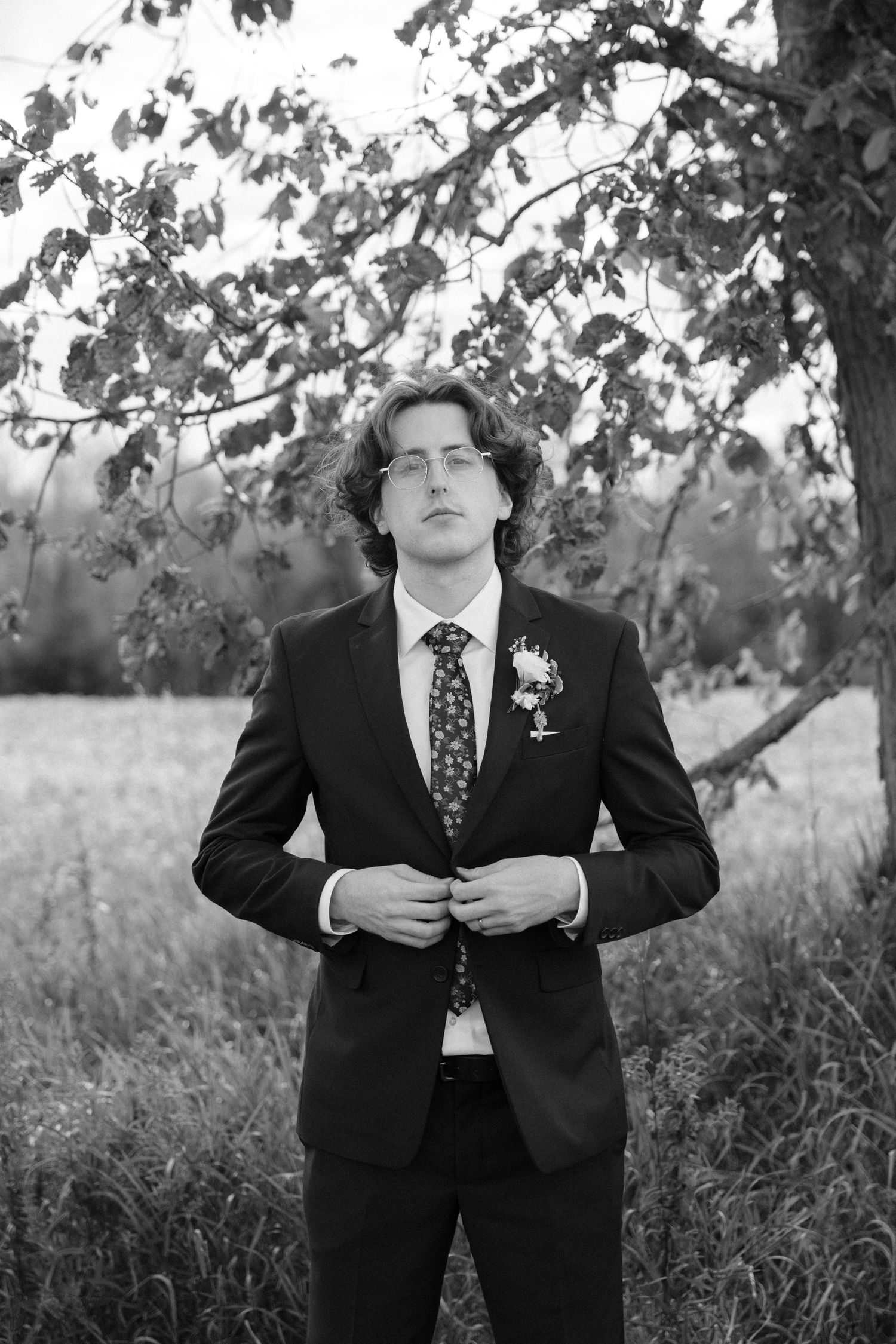 Black and white portrait of a dapper groom in a suit and floral tie standing beneath a tree outdoors.