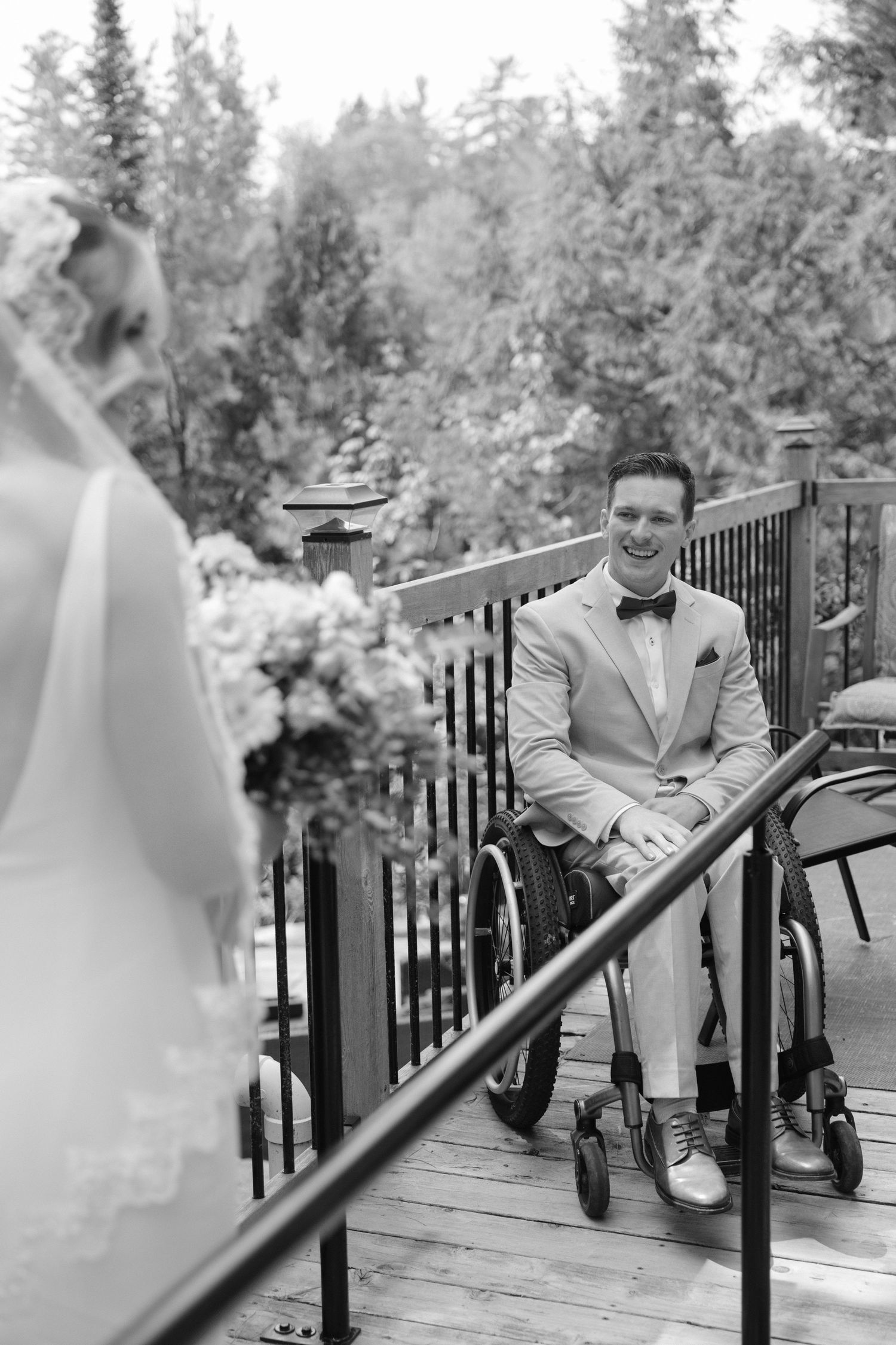 Groom in wheelchair smiling at bride during outdoor wedding ceremony on wooden deck.