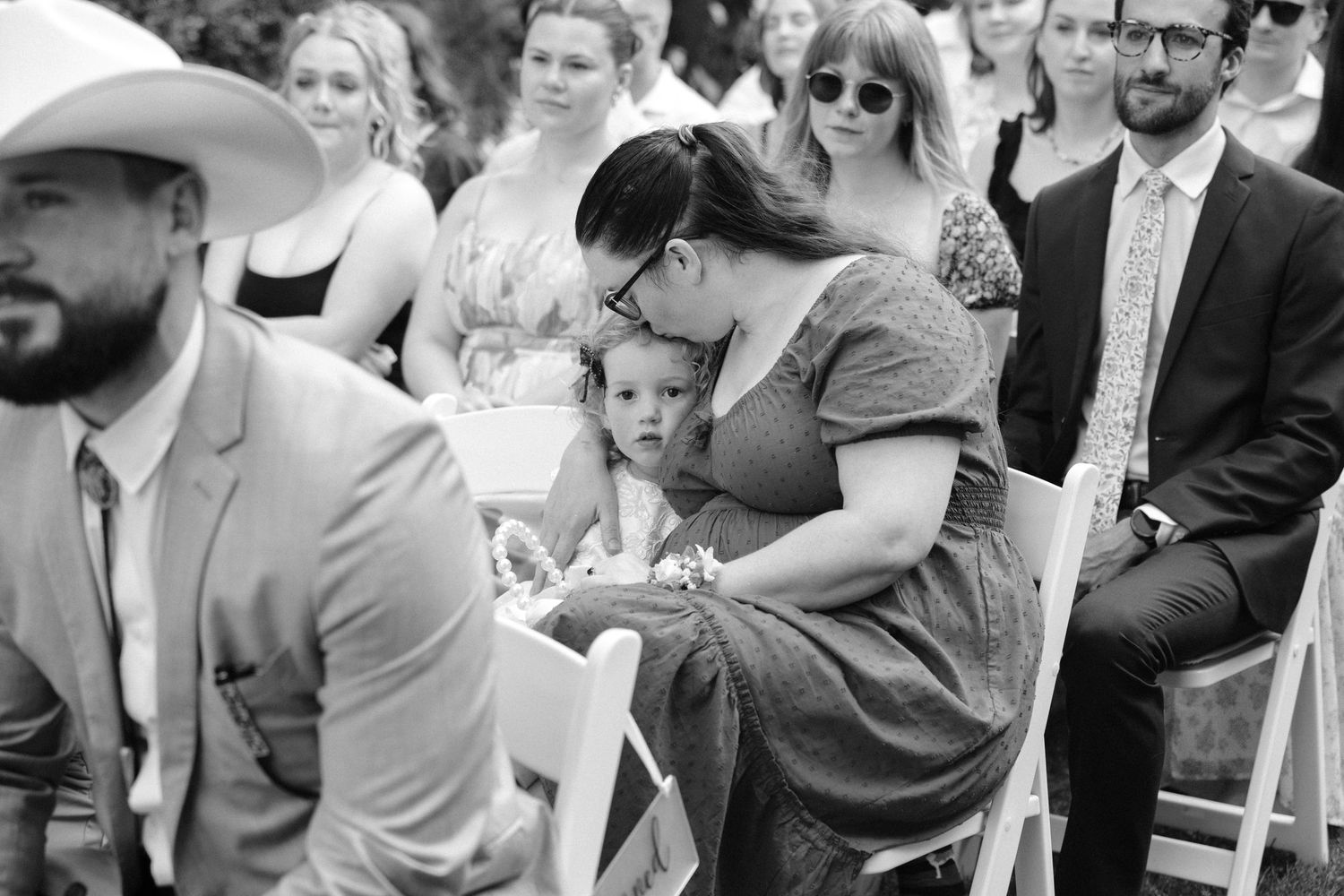 Black and white photo of wedding guests seated outdoors with mother comforting young child.