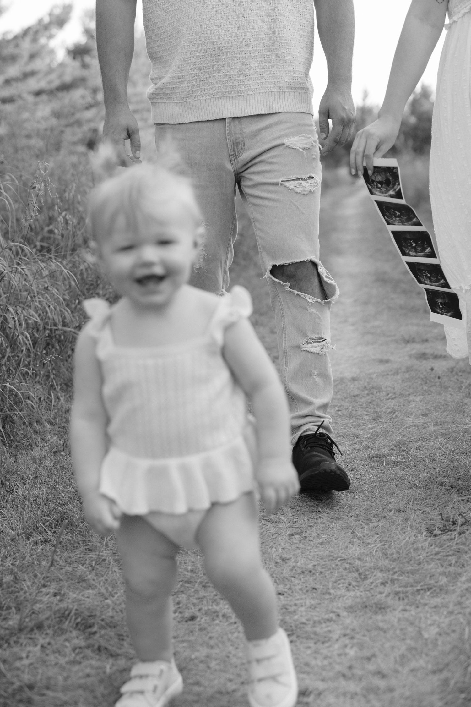 Black and white photo of a laughing toddler walking ahead of parents holding ultrasound images.