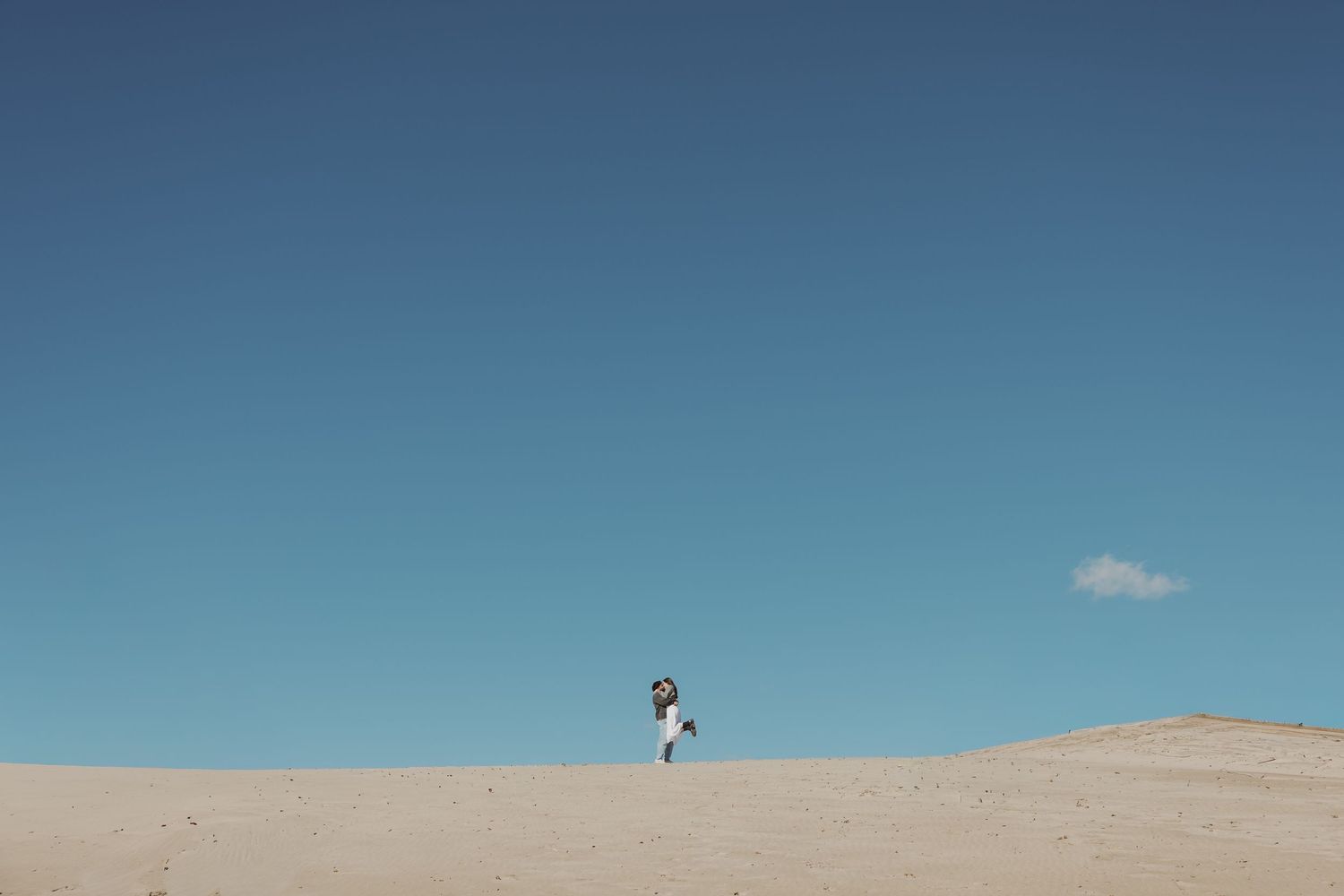 Couple embracing on a vast sandy dune under a clear blue sky in a wide landscape shot.
