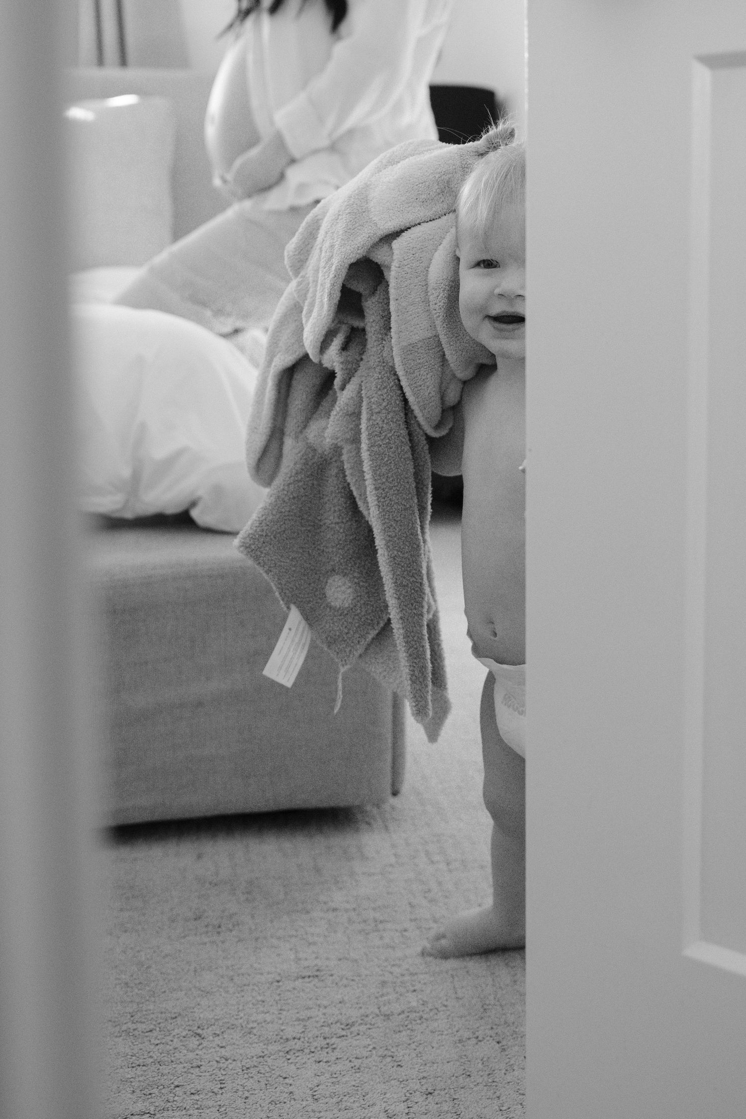 Black and white photo of a laughing baby wrapped in a towel standing in a bedroom doorway.