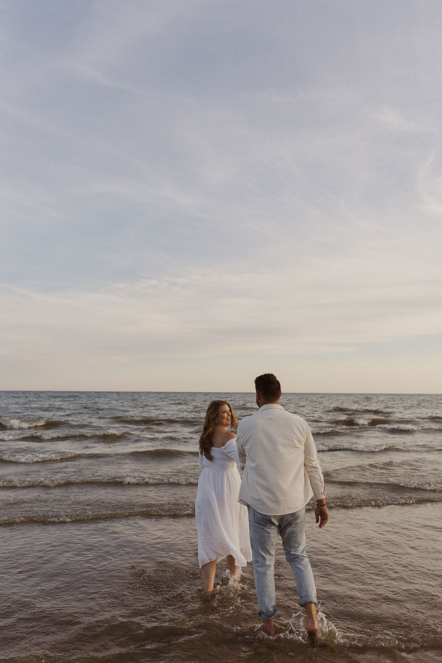 Couple walking hand in hand through ocean waves on a beautiful beach at dusk.