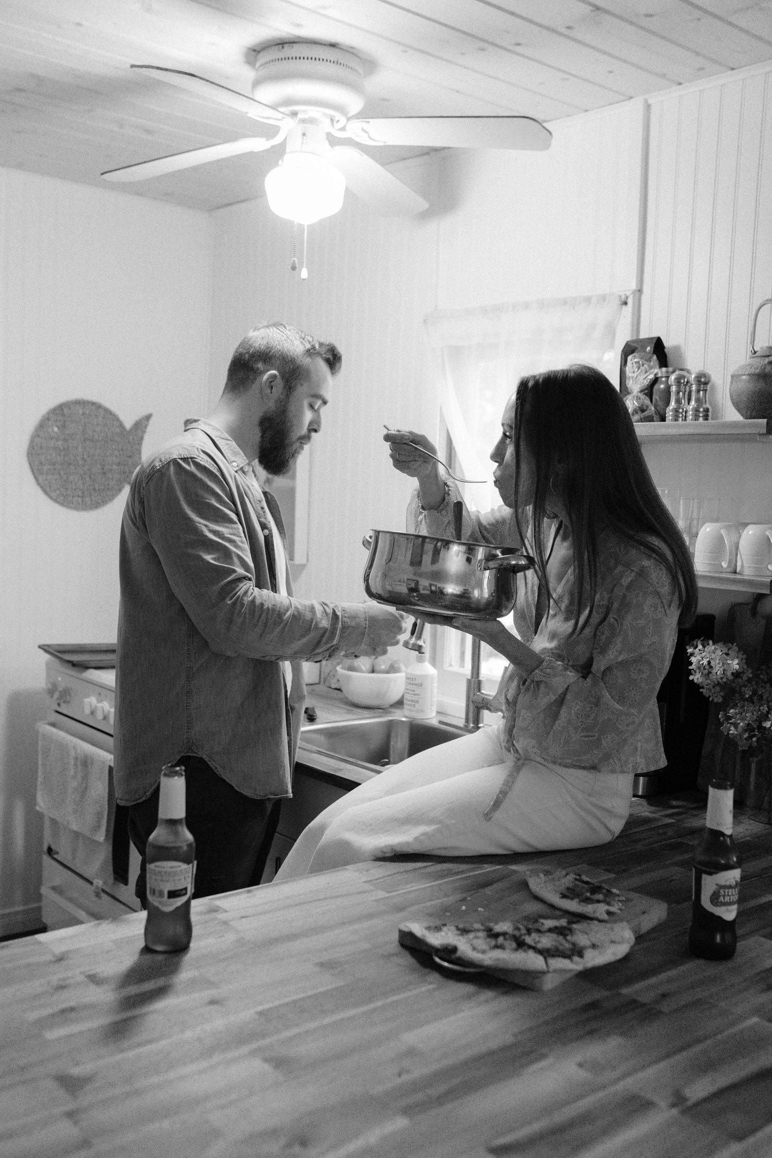 Black and white photo of bearded man feeding woman sitting on kitchen counter indoors.