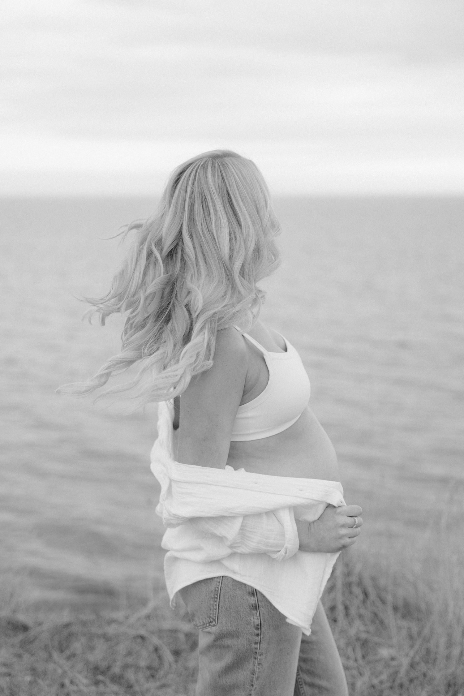 Black and white photo of pregnant blonde woman facing ocean with wind-blown hair.