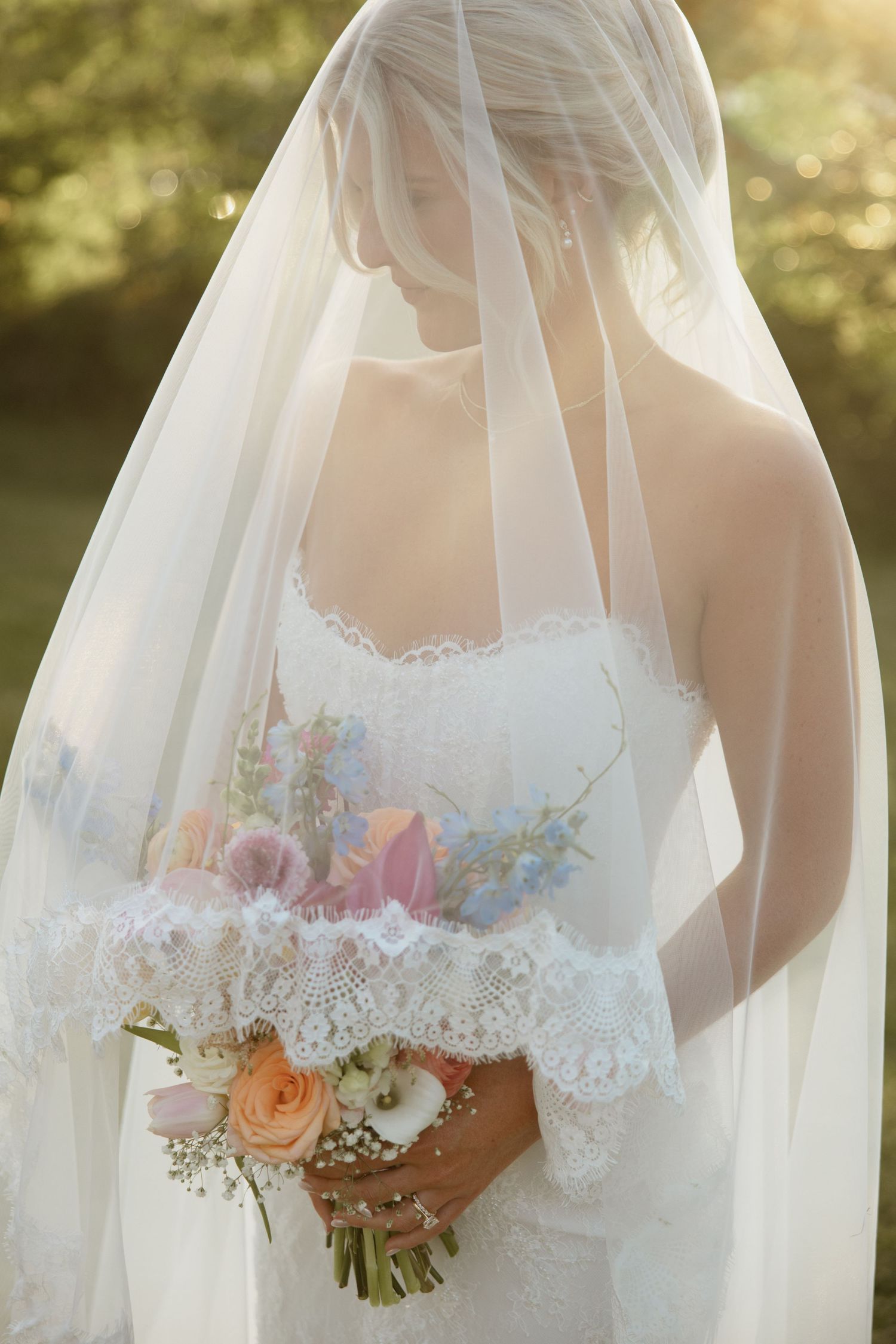 Bride in white lace gown and flowing veil holds pastel bouquet of roses outdoors in soft golden light.