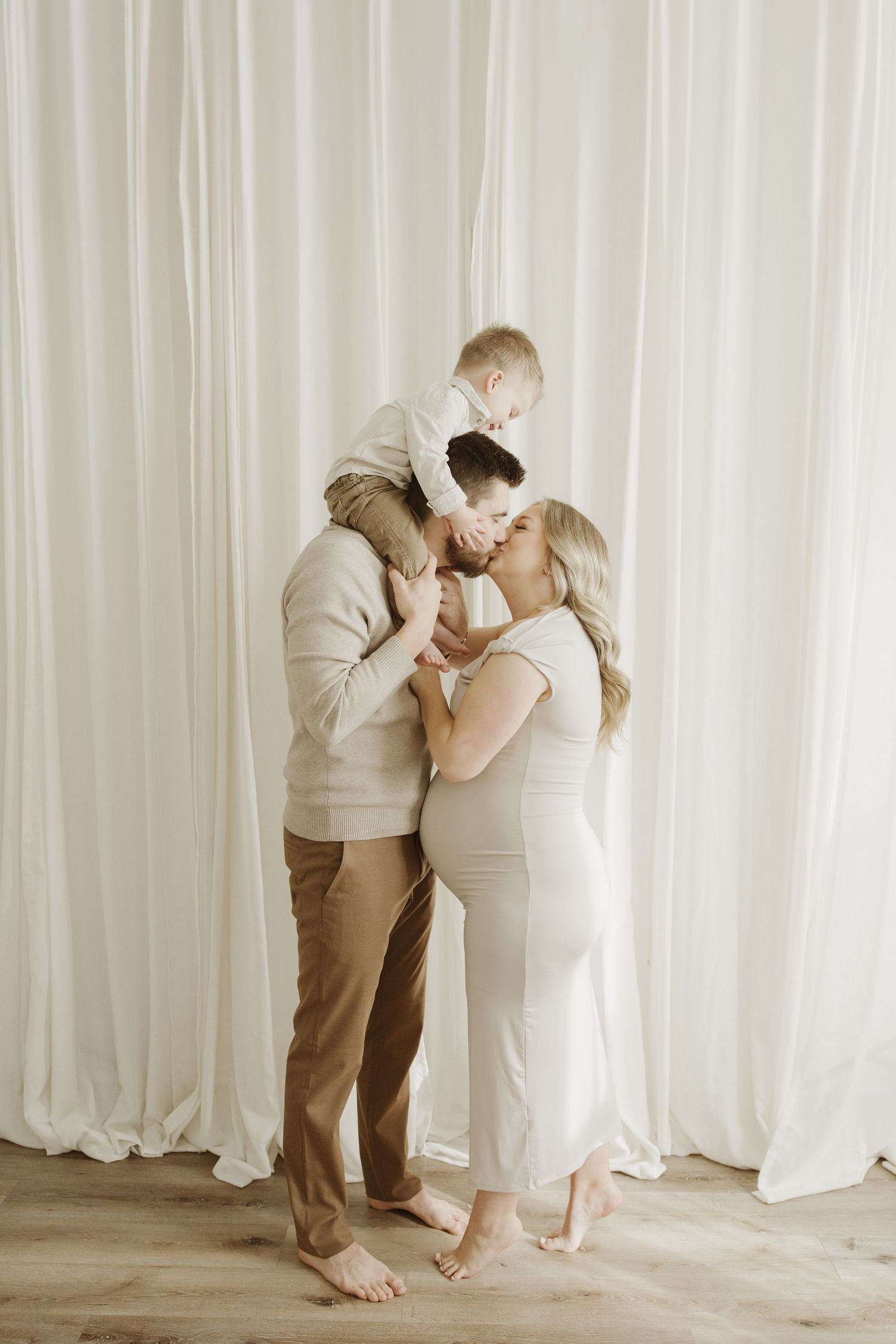 Happy family with pregnant mother in white dress and father holding baby on shoulders against white curtains.