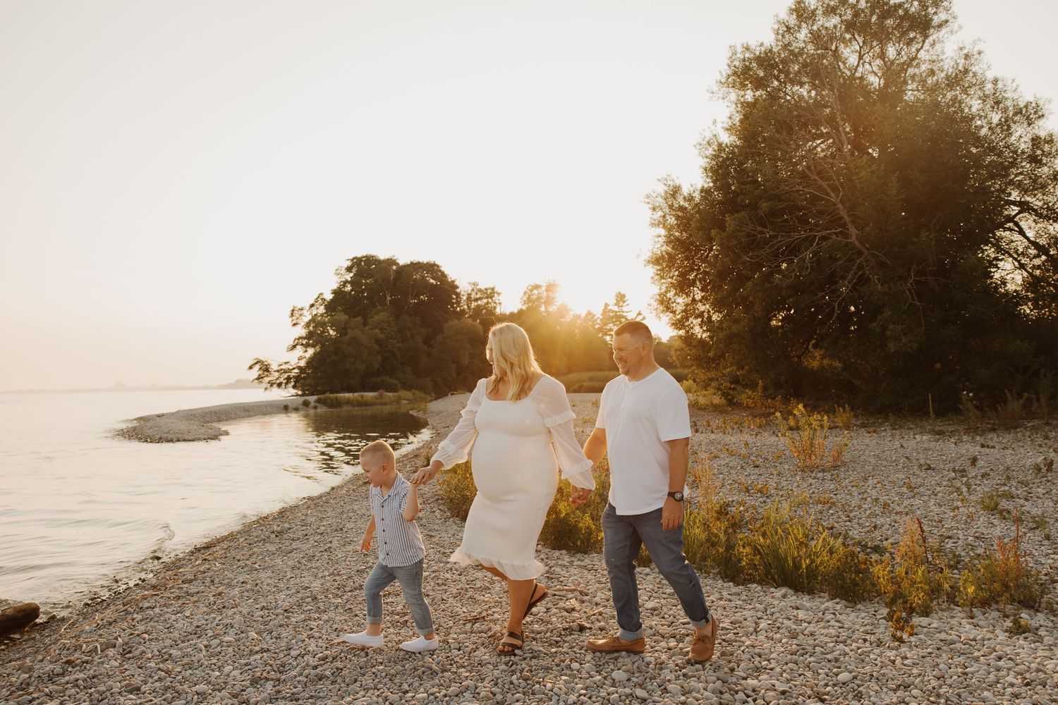 Pregnant woman in white dress holds hands with partner and child walking along a rocky lakeside beach at sunset.