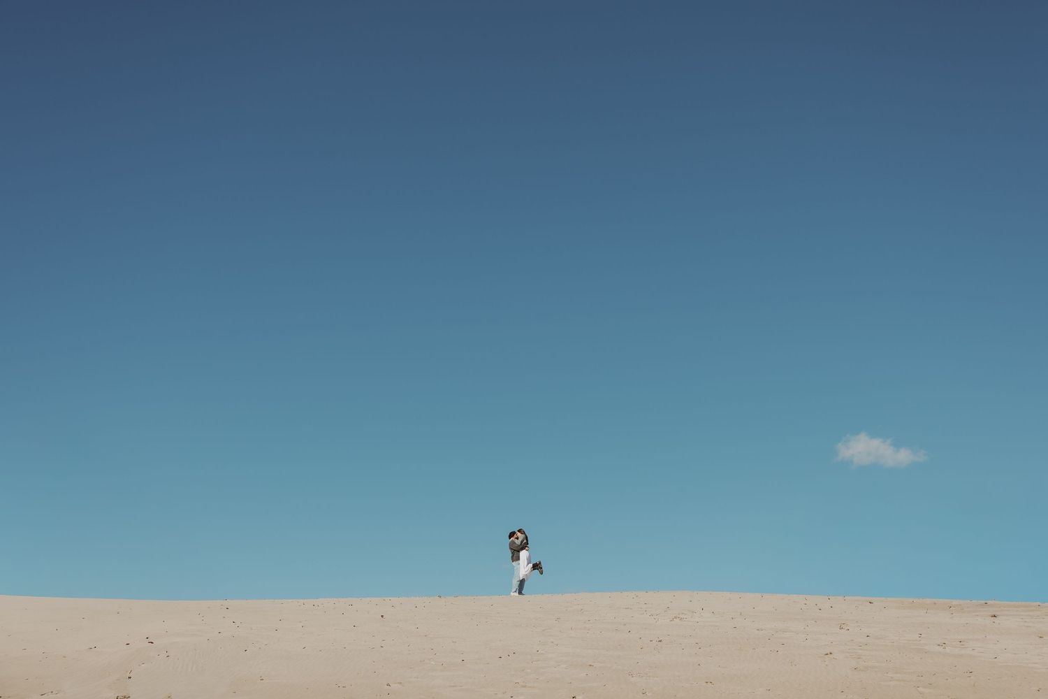 Couple embracing on sandy dunes under vast blue sky in minimalist landscape.