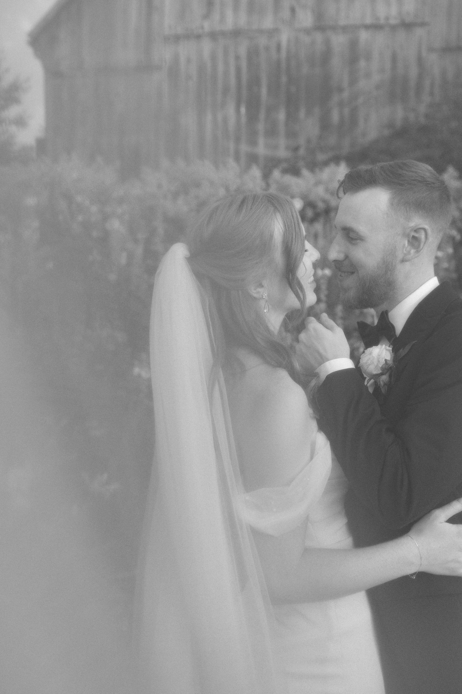 Black and white dreamy double exposure photo of smiling groom gazing at bride in veil.