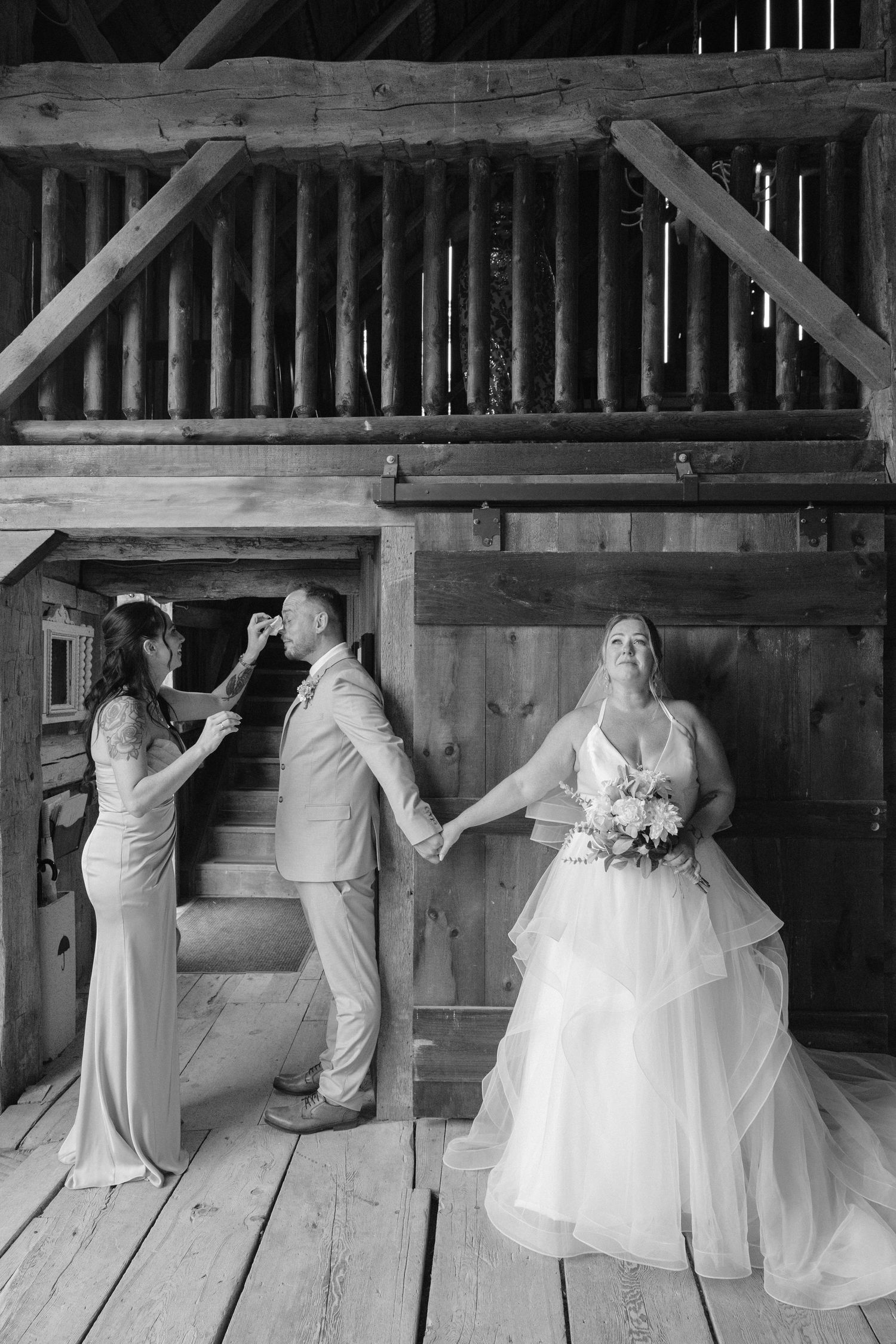 Black and white photo of bride and groom holding hands in rustic barn while bridesmaid adjusts grooms boutonniere.