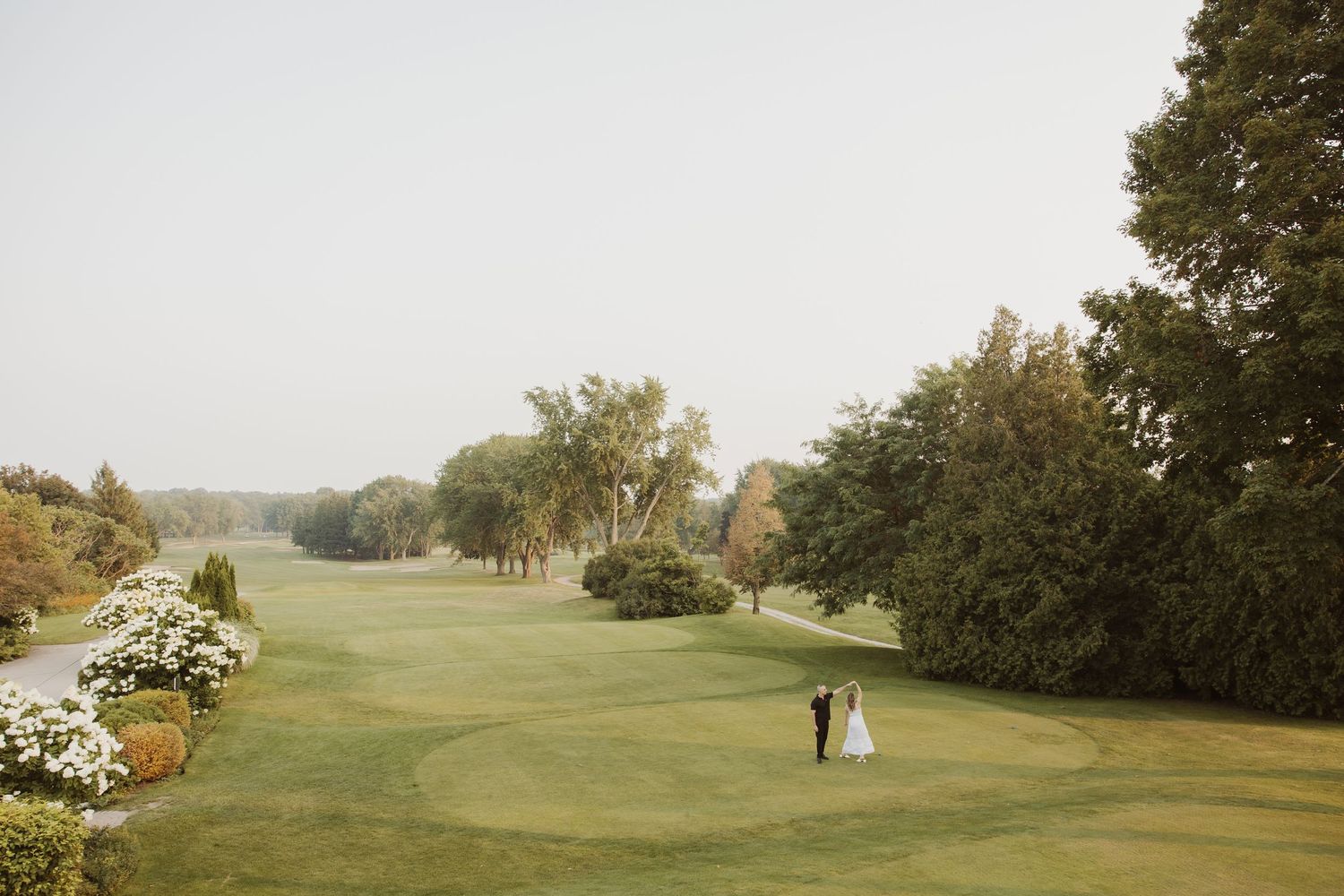 Bride and groom standing together on a vast green golf course surrounded by lush trees on a misty day.