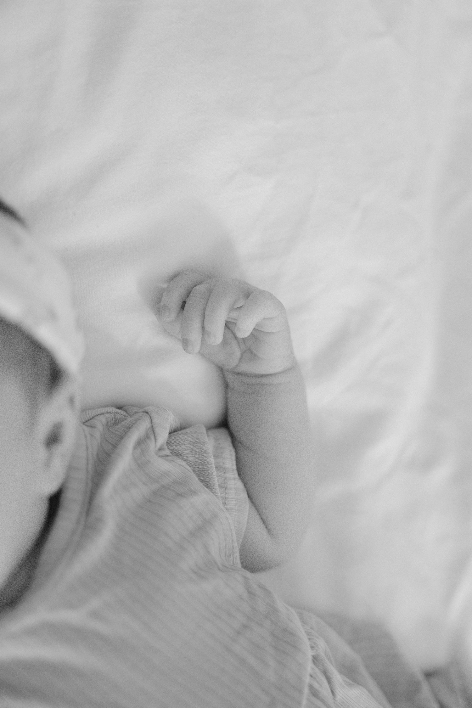 Black and white close-up of a sleeping newborn baby's tiny fist on white bedding.