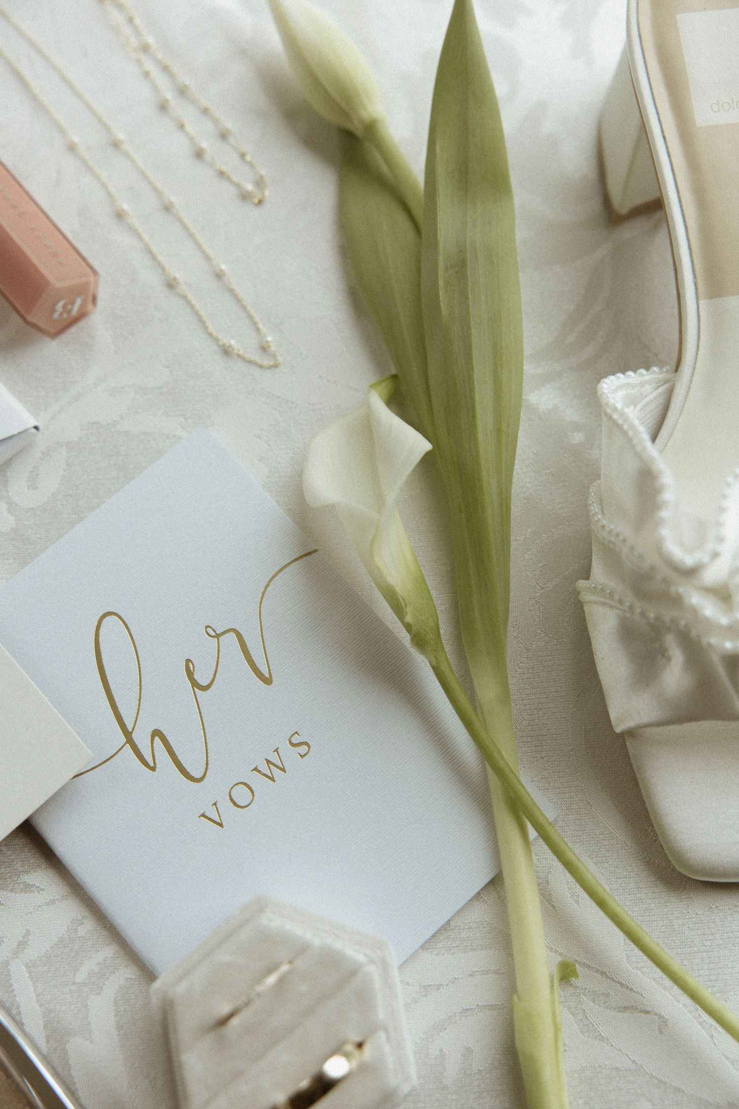 Elegant bridal flat lay with her vows book, white tulips, pearl heels, gold necklace, and nude lipstick on ivory fabric.