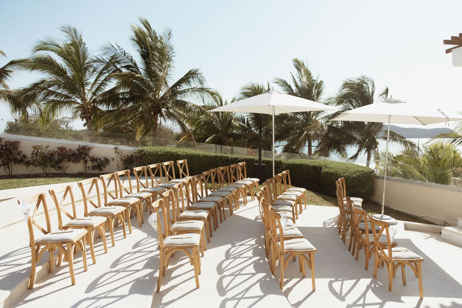 Outdoor wedding ceremony setup with wooden chairs arranged on a white terrace surrounded by tropical palm trees.