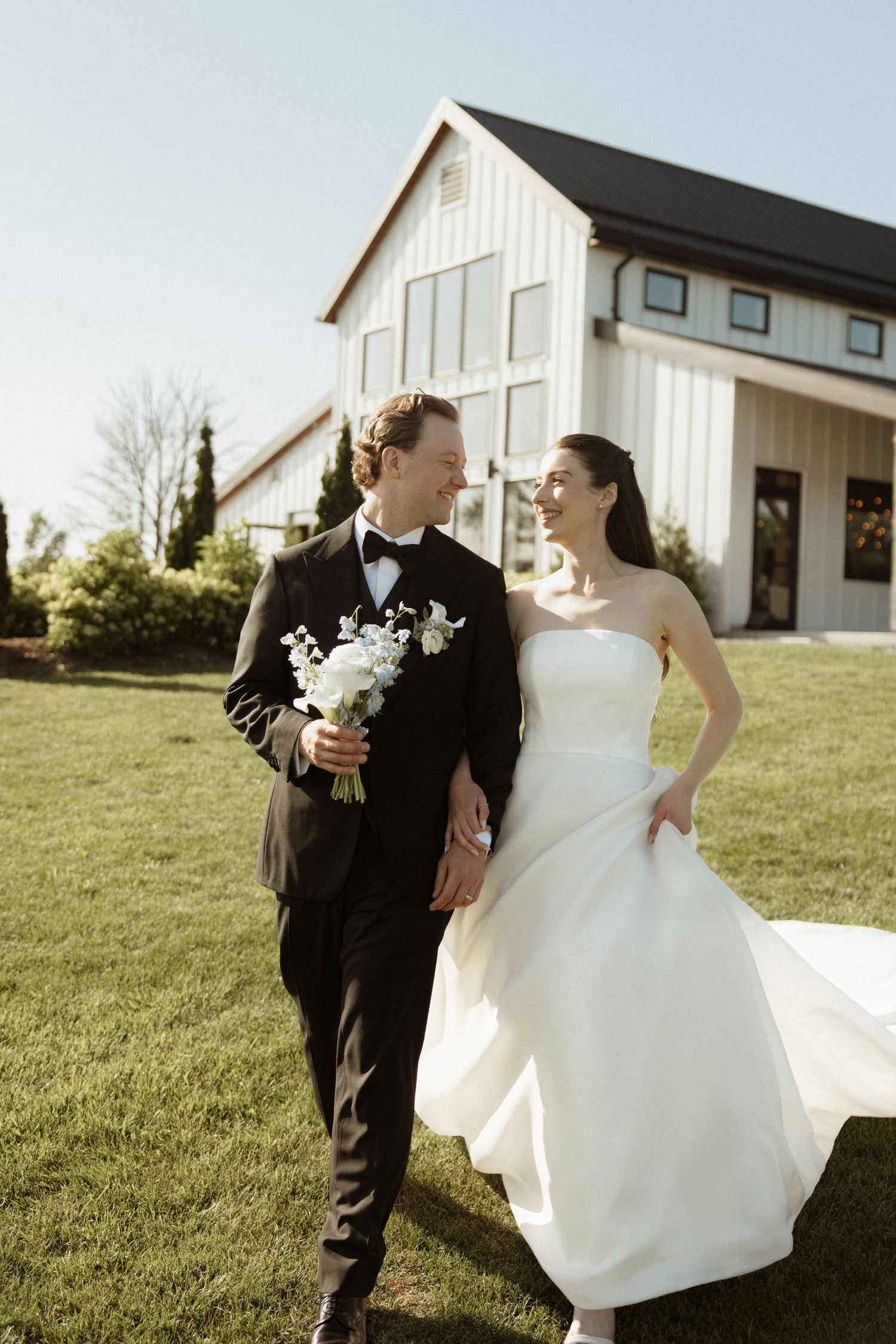 Bride and groom walking on green lawn in front of white barn venue, holding bouquet on sunny wedding day.